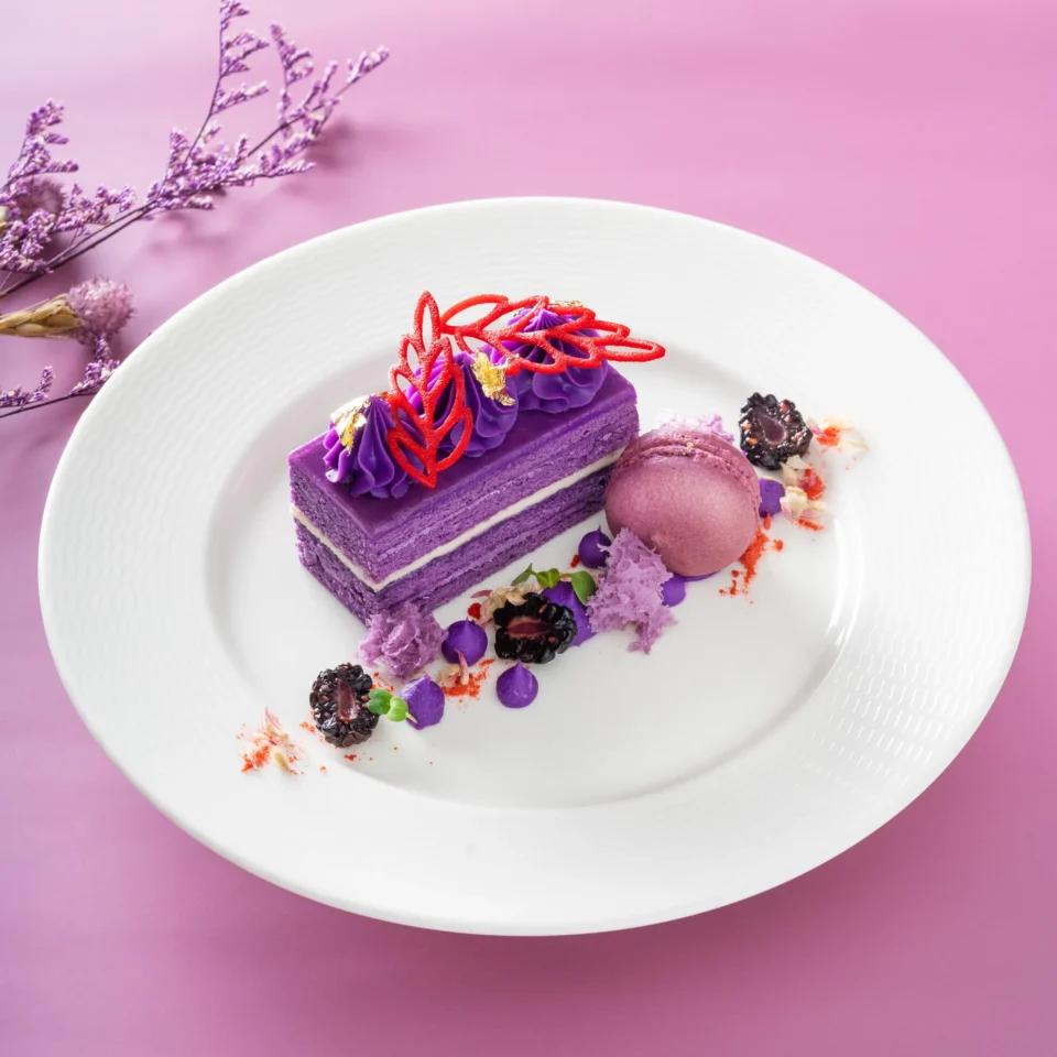 Purple layered cake slice with purple frosting and red lattice topping on a white textured plate against a pink background.