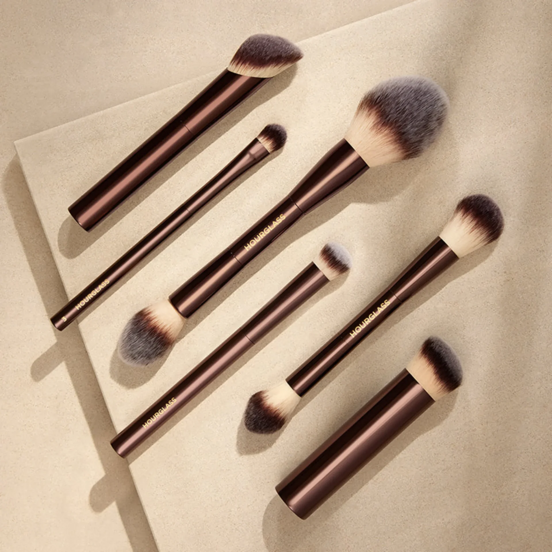 Set of brown-handled makeup brushes with fluffy bristles arranged on a beige surface.