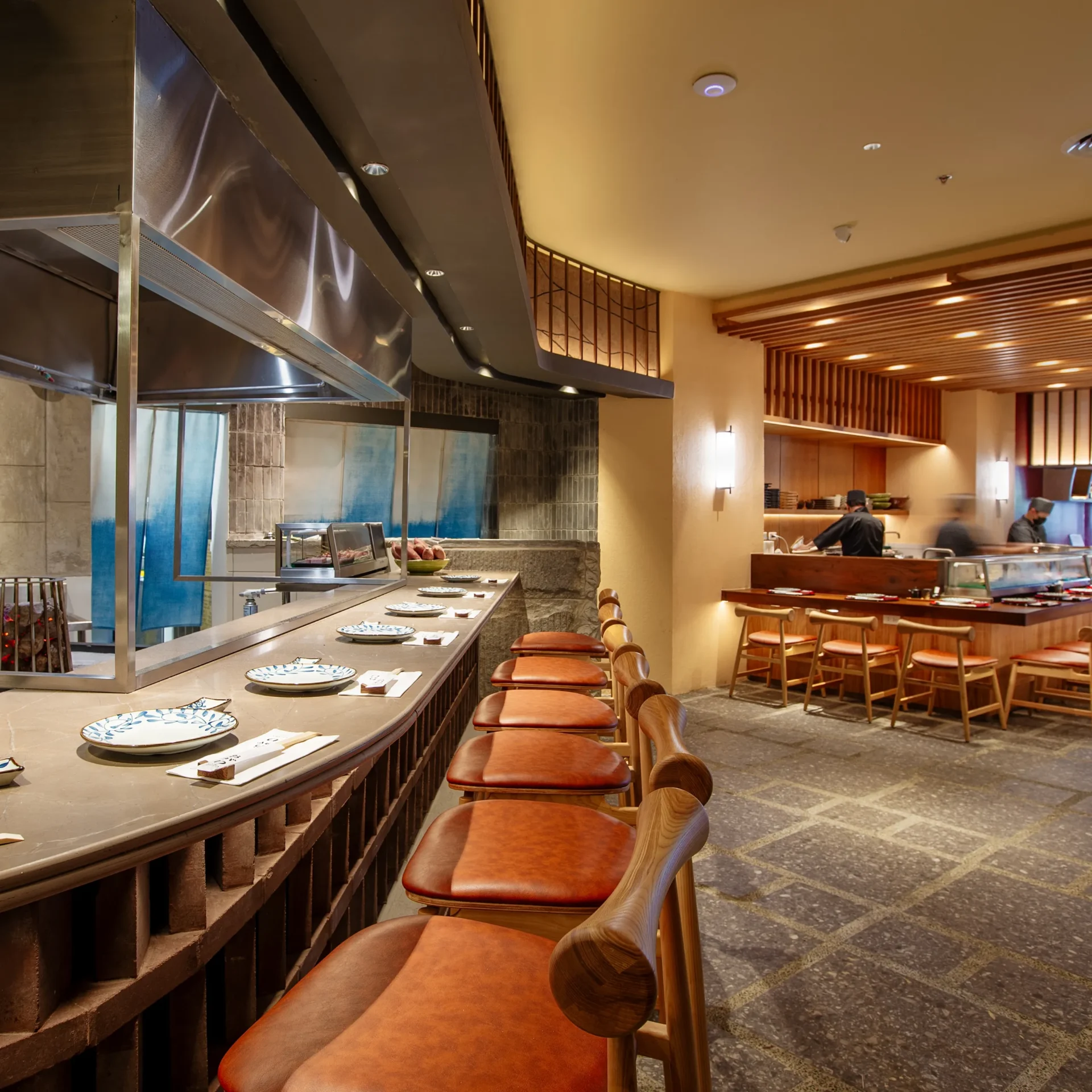Bar-style restaurant counter with a row of brown leather stools, plates set along the counter, and a busy kitchen area with stainless steel equipment in the background.
