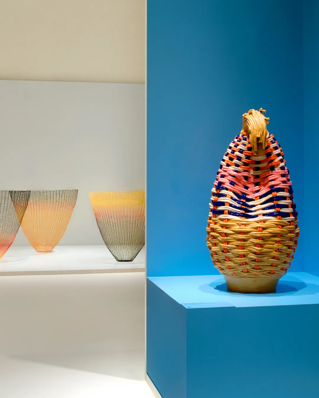 Tall multicolored woven basket sculpture on a blue pedestal in a gallery, with three wire-mesh vases in the background on the left.