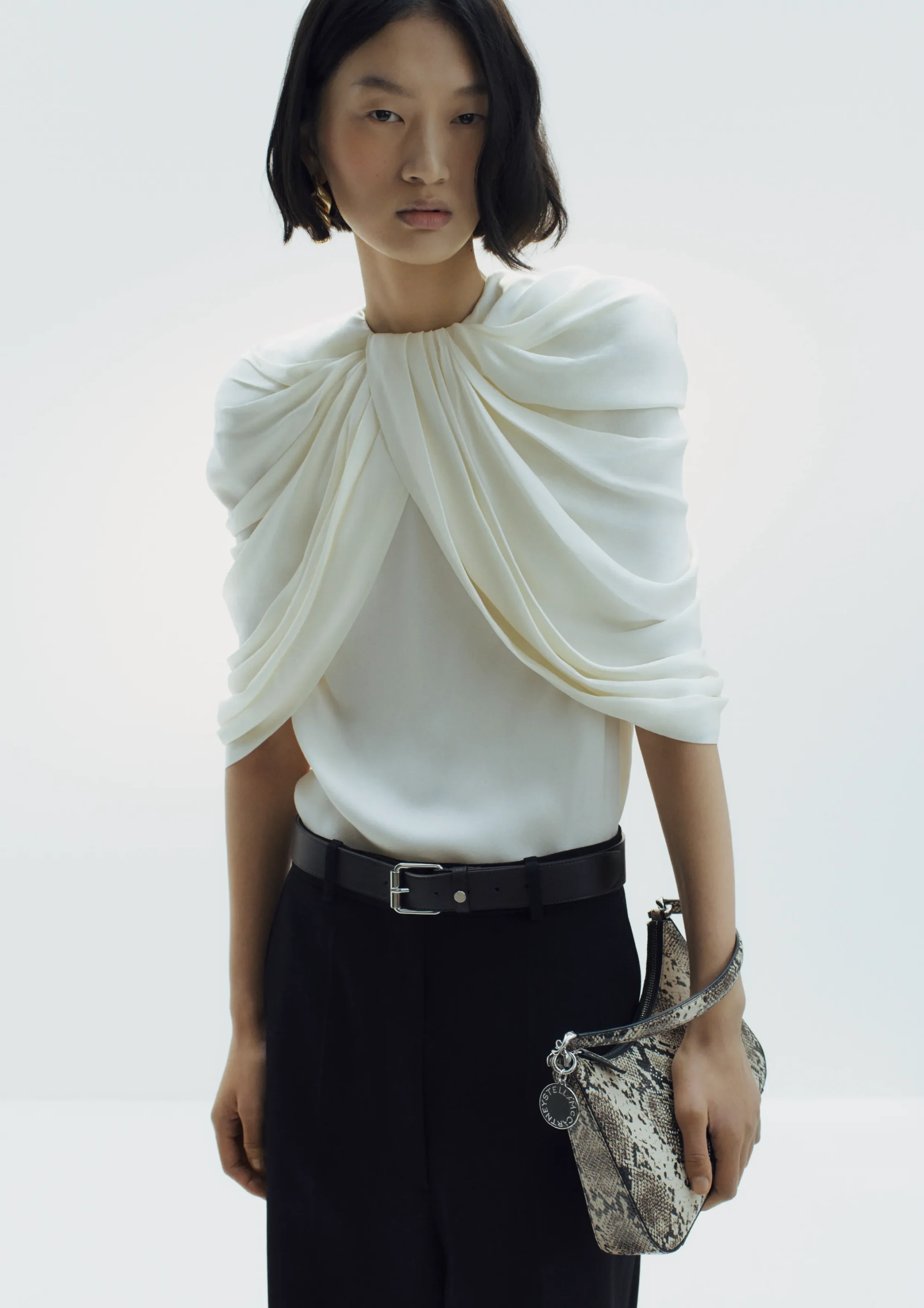 Model wearing a white draped top with black trousers and a snakeskin handbag held at her side in a minimalist setting.