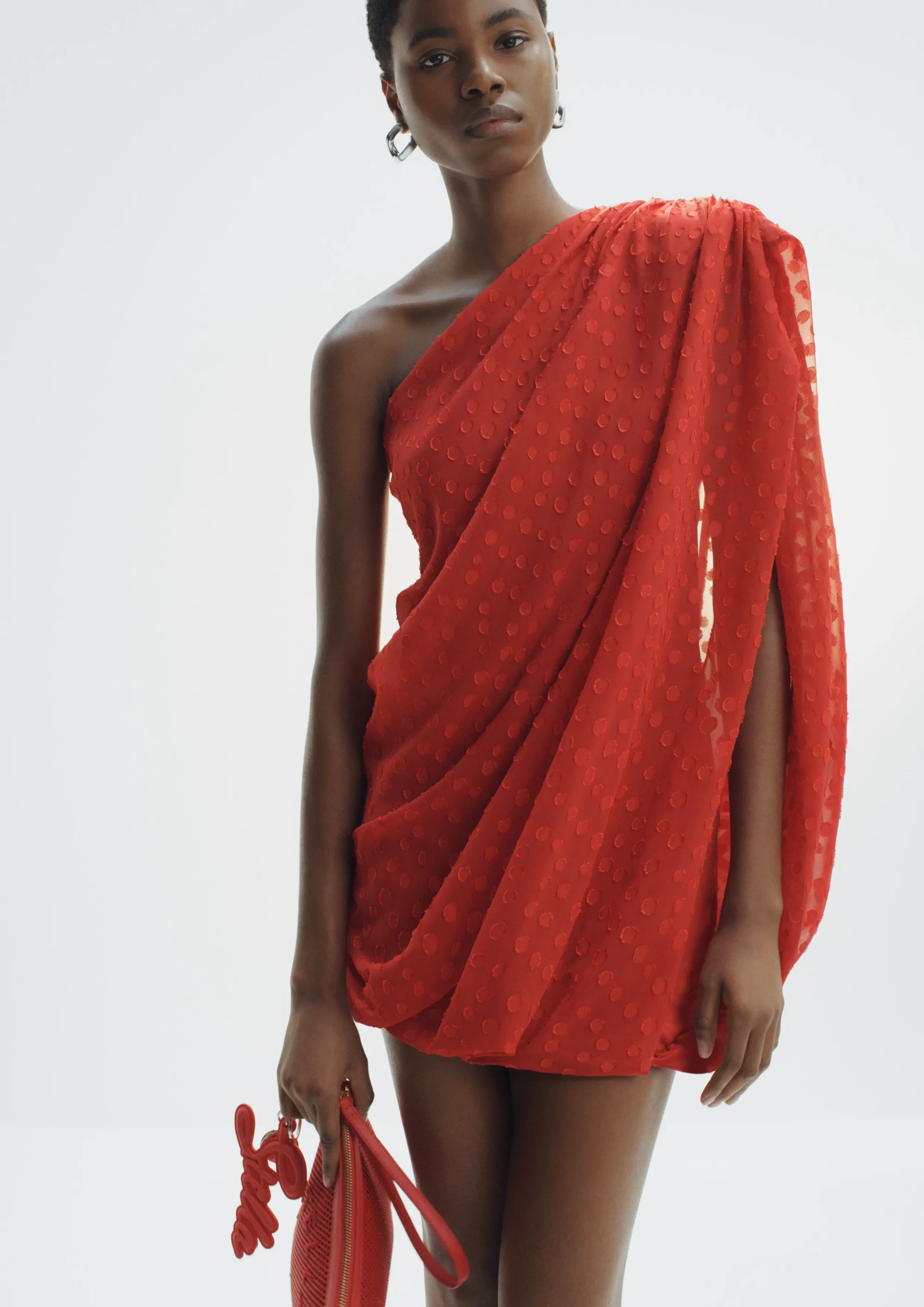 Model wearing a red one-shoulder dress with a dotted texture, holding a matching red handbag against a light background.