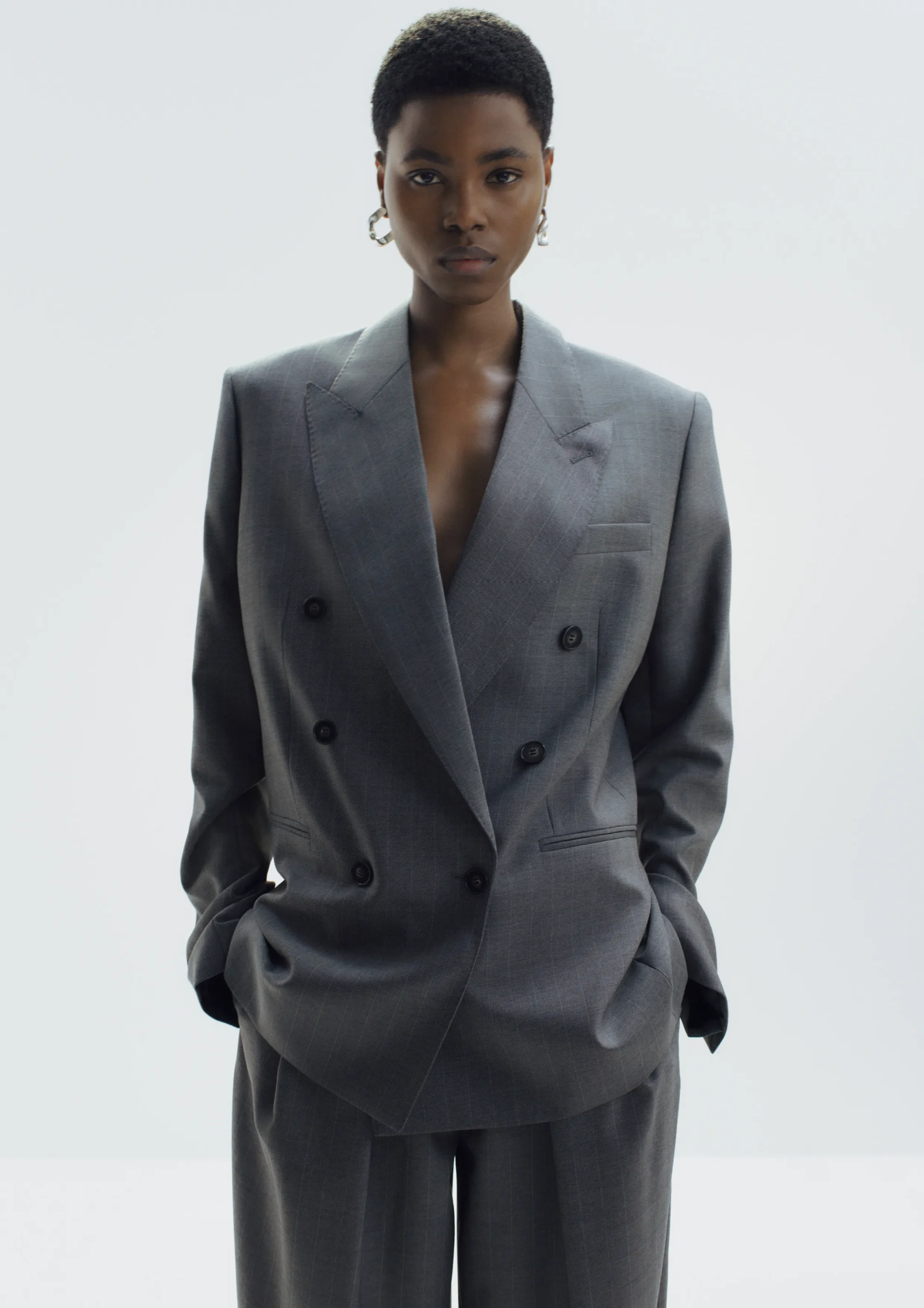 Professional portrait of a Black woman in a gray double-breasted suit, standing against a light backdrop, hands in pockets.