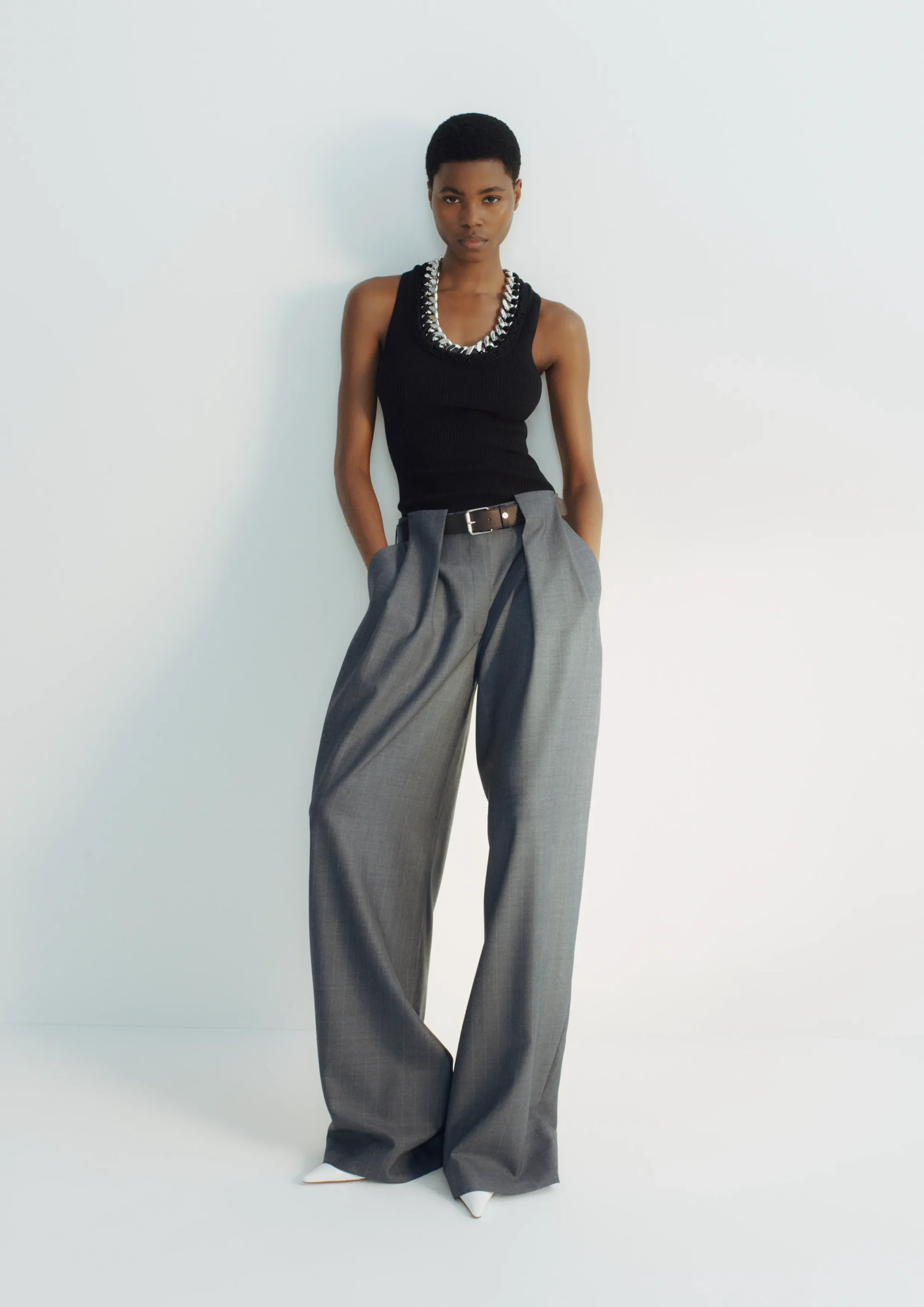 Model wearing a black sleeveless top with a chunky silver necklace and wide-leg gray trousers, standing against a pale wall.