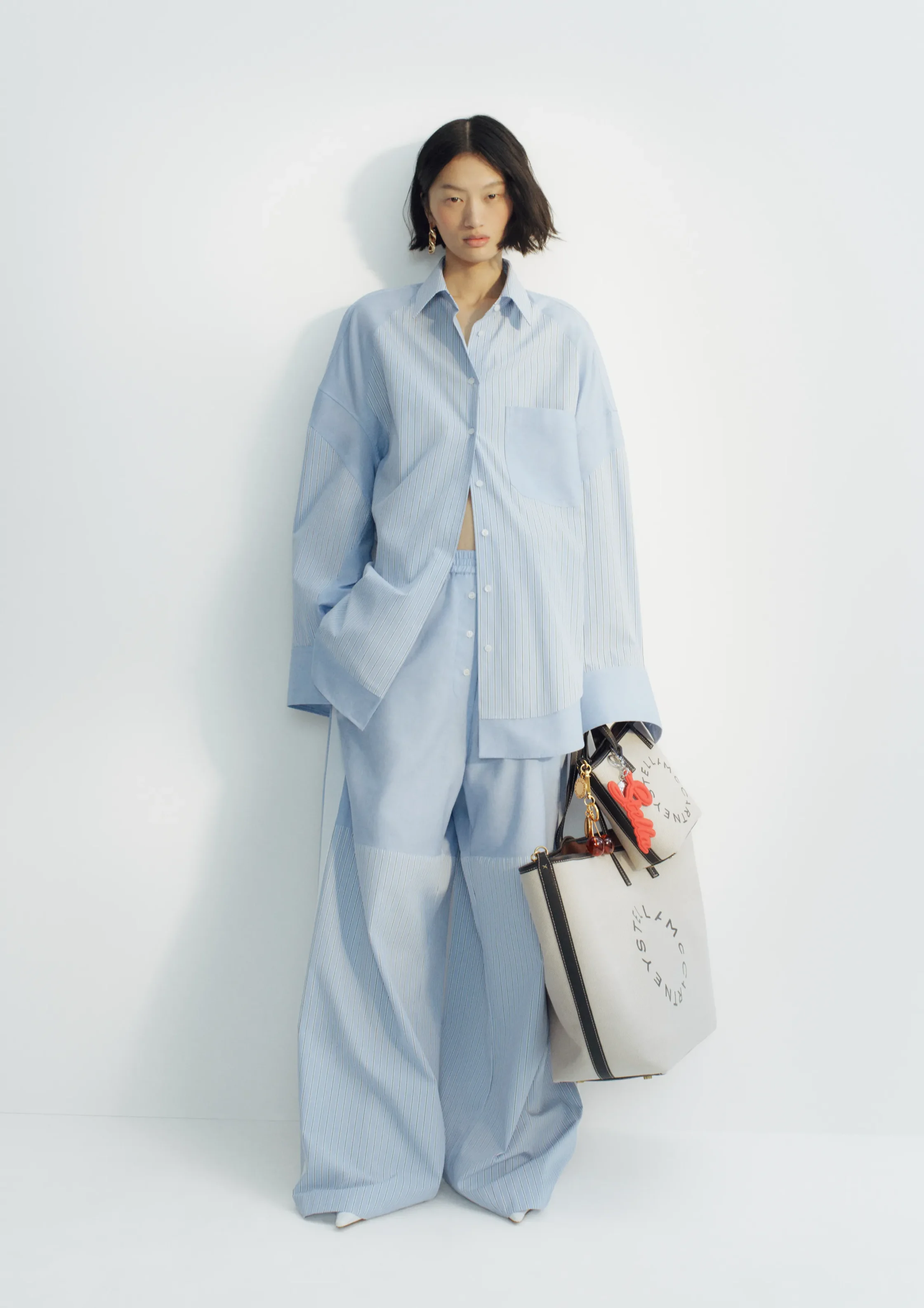 Person wearing an oversized light blue striped shirt and wide-leg pants, standing against a pale background while holding a white tote bag with black straps and red accents