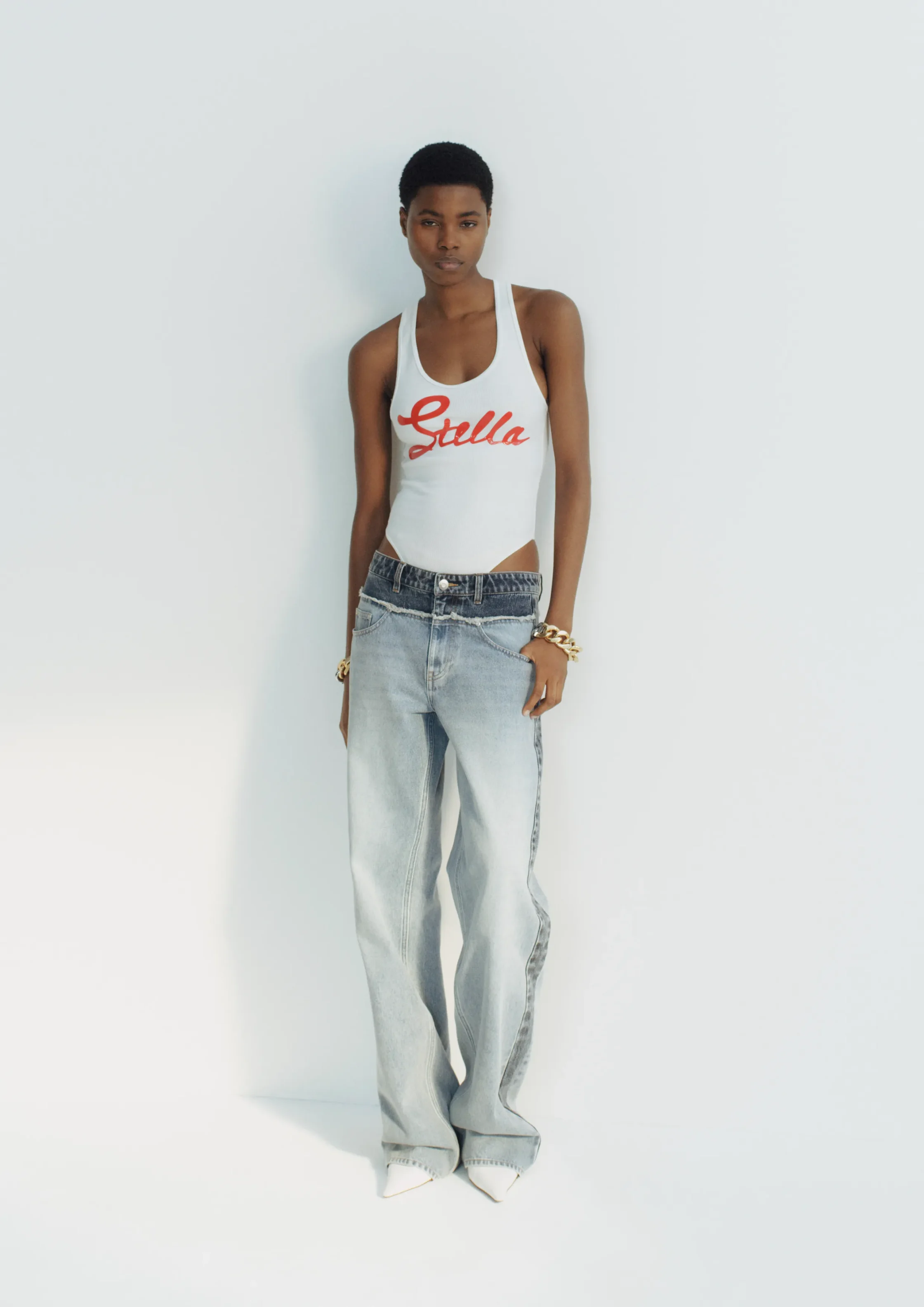 Model wearing a white tank top with red 'Stella' script, light-wash jeans, and gold bracelets, standing against a pale backdrop.