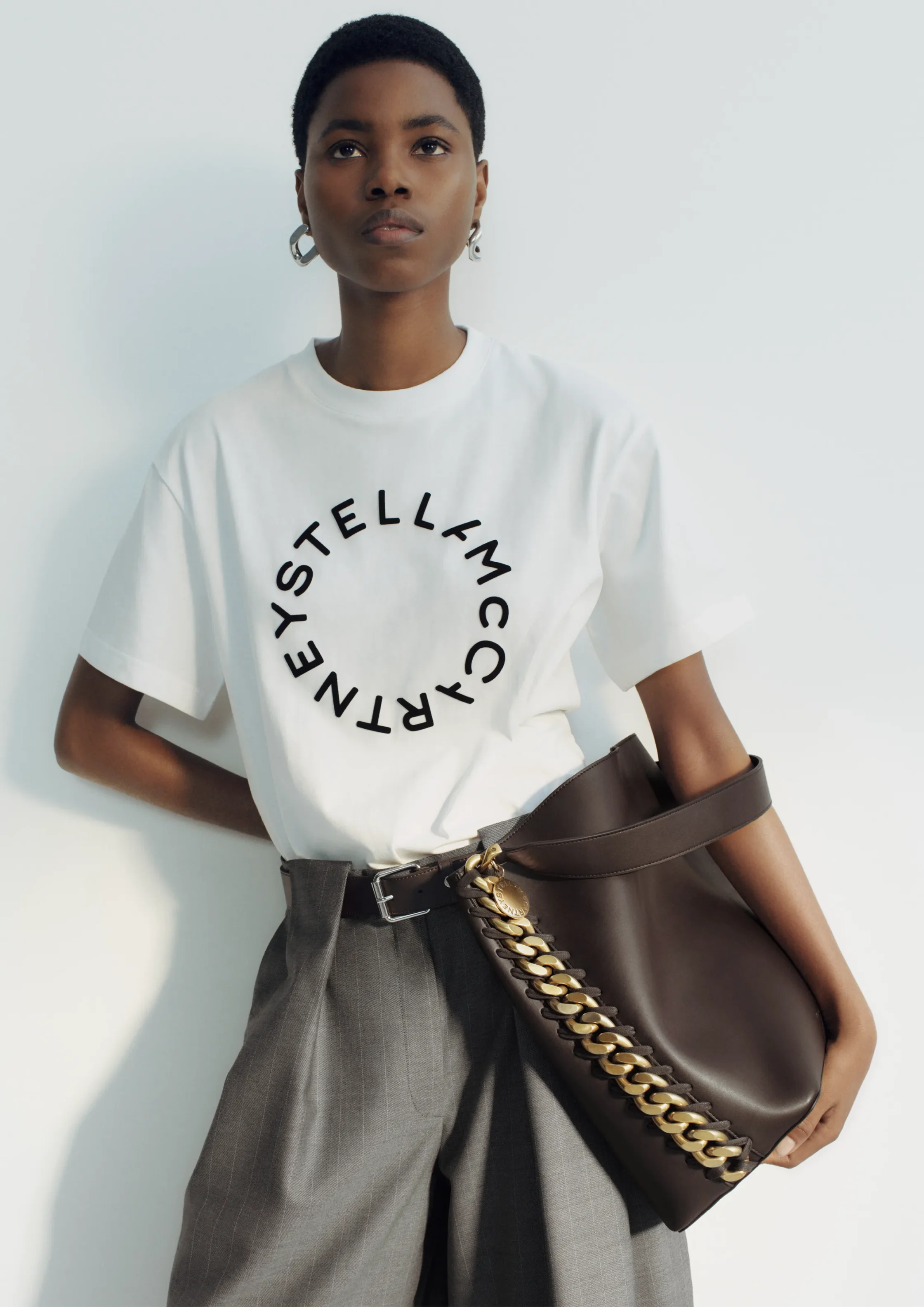 Portrait of a Black woman with short hair wearing a white t-shirt featuring a circular Stella McCartney logo, paired with gray pinstripe trousers and a brown handbag with a chunky gold chain over the shoulder.