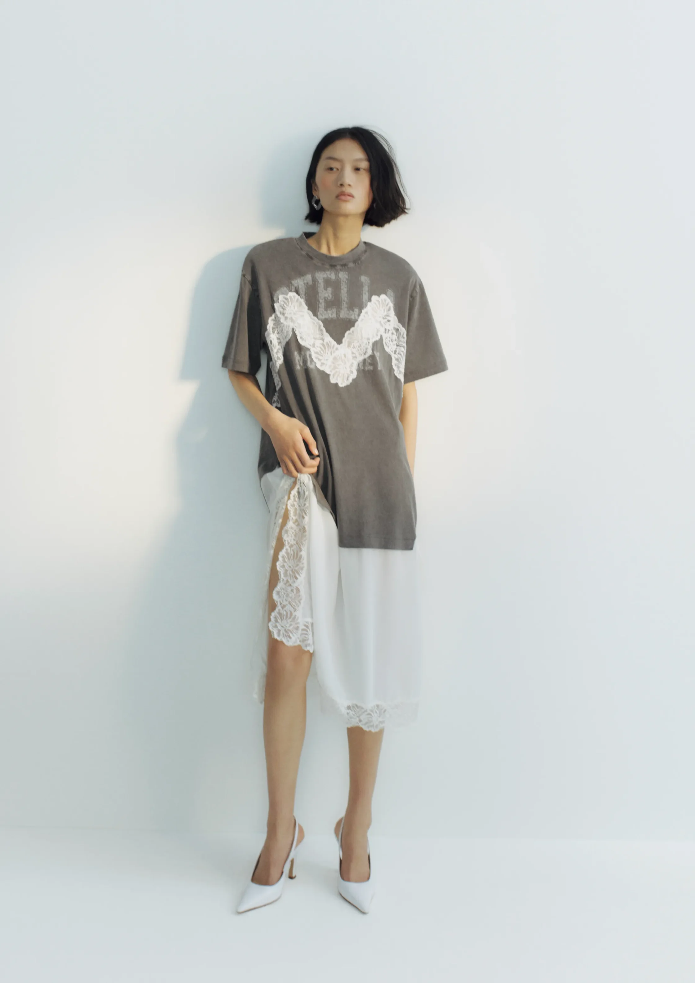 Full-body fashion shot of a woman standing against a pale wall, wearing an oversized gray T-shirt with white lace detailing and a white sheer skirt with lace trim, and white heels, looking off to the side