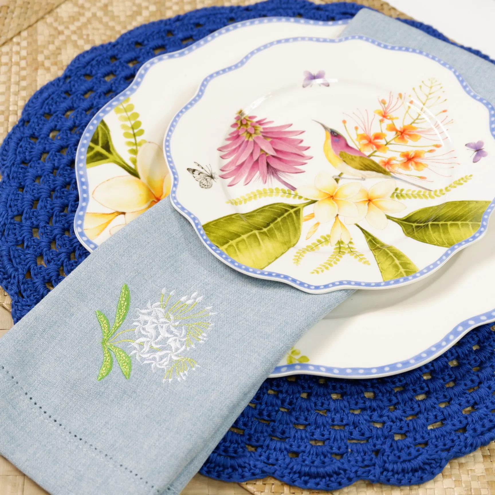 Decorative porcelain plate with pink flower and a small bird, placed on a blue crocheted placemat with a light blue napkin nearby. casa juan philippine handsome sunbird collection