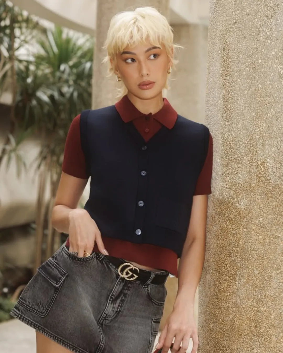 Person with short blonde hair wearing a burgundy polo and navy vest, denim mini skirt with a Gucci belt, leaning against a textured column outdoors.
