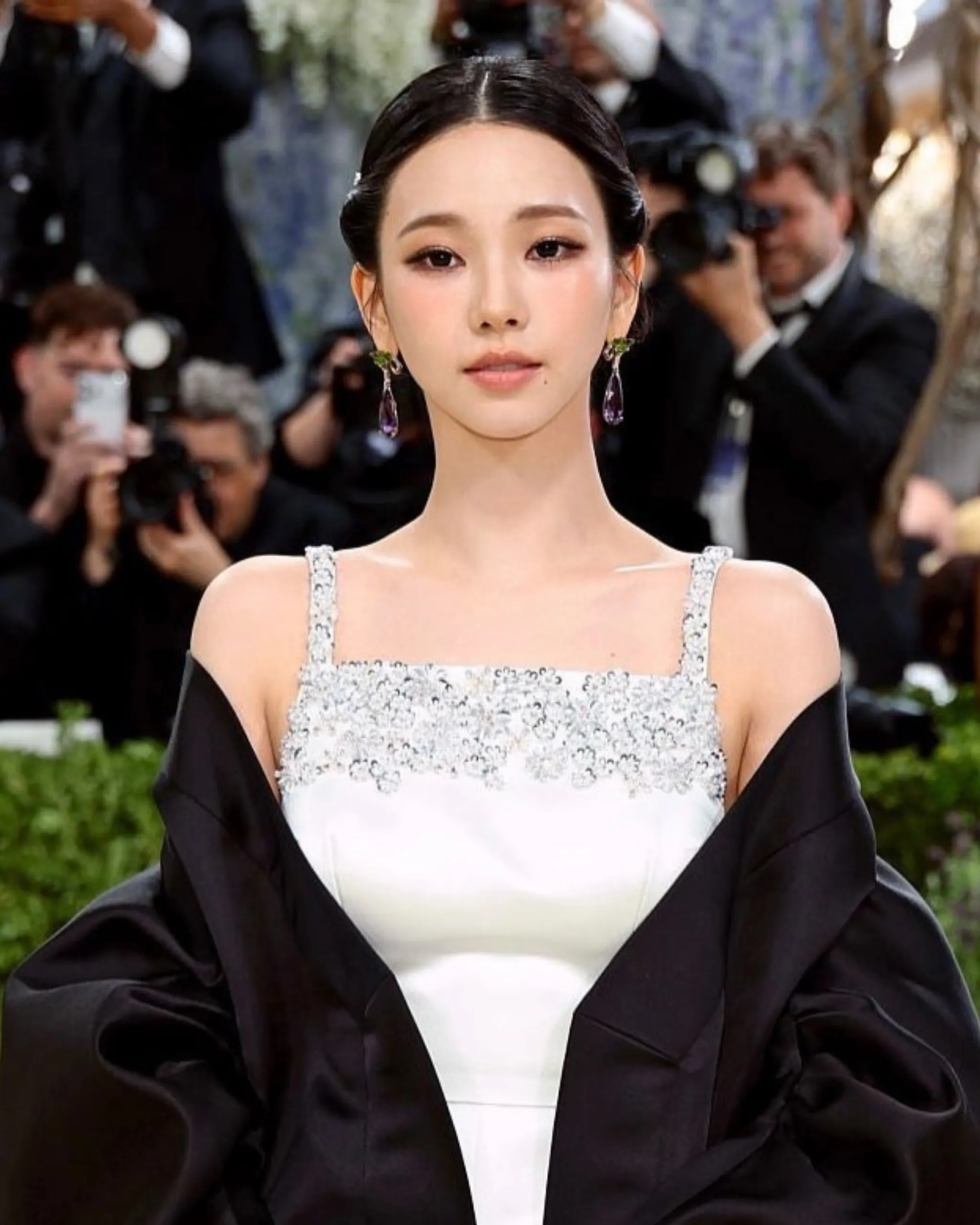 Woman on a red carpet wearing a silver-embellished white gown with a black shawl; photographers in the background.