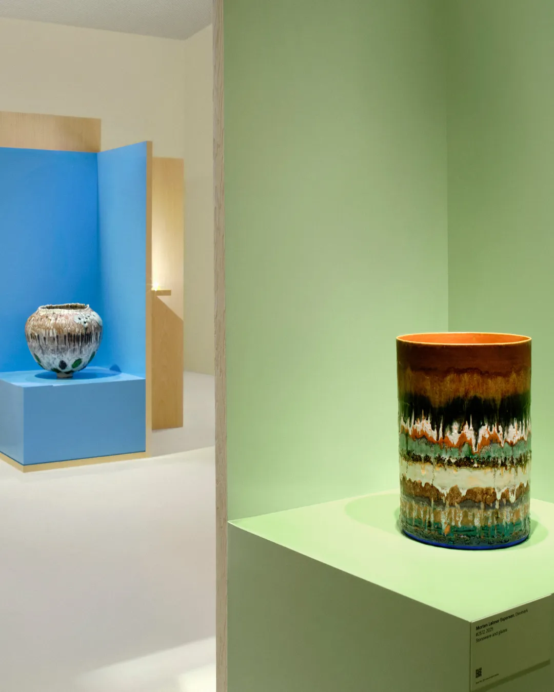 Multicolored ceramic vase on a lime pedestal in a gallery, with another blue display alcove in the background.