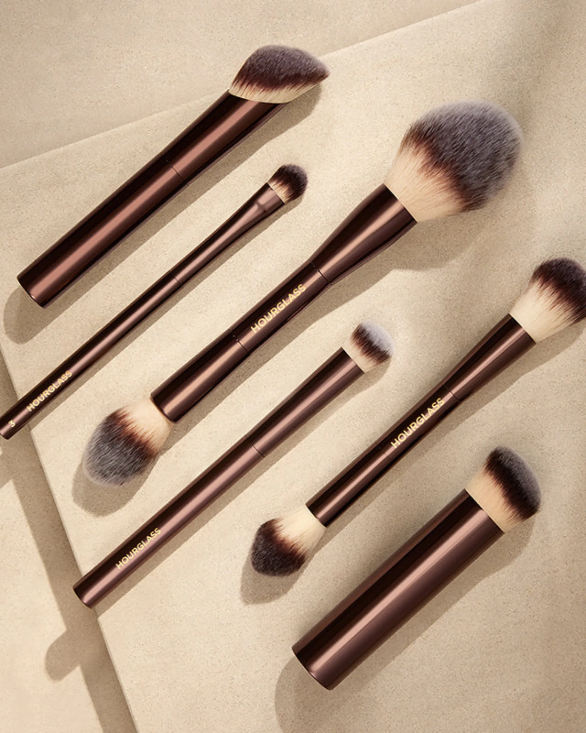 Several makeup brushes with brown handles and cream bristles laid diagonally on a beige surface.