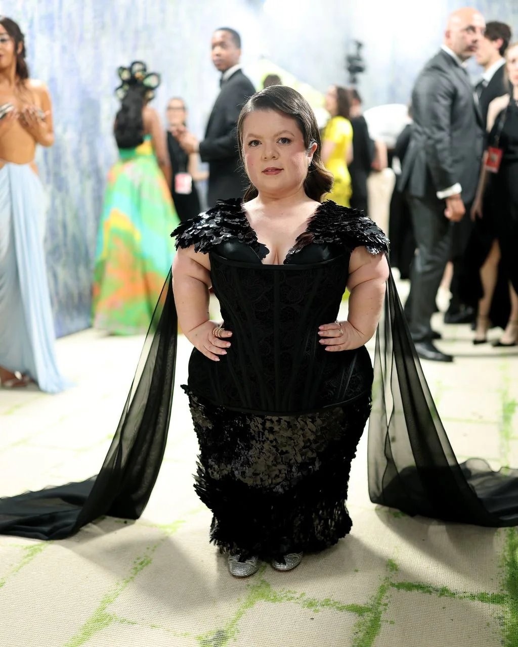 Woman in a black lace gown with feathered shoulders posing with hands on hips at a formal event.