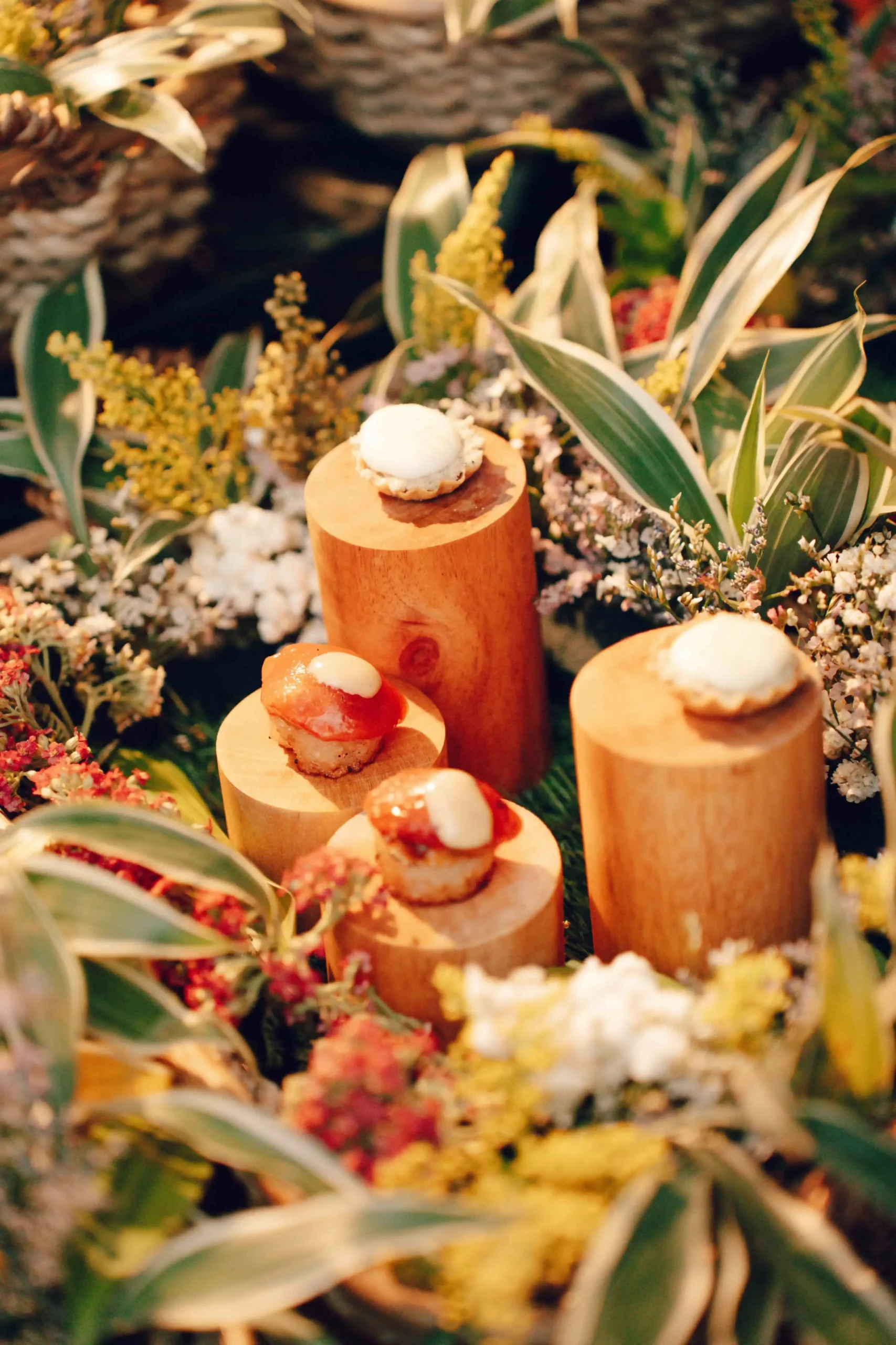 Small pastries topped with white cream on wooden rounds among greenery in a garden display.