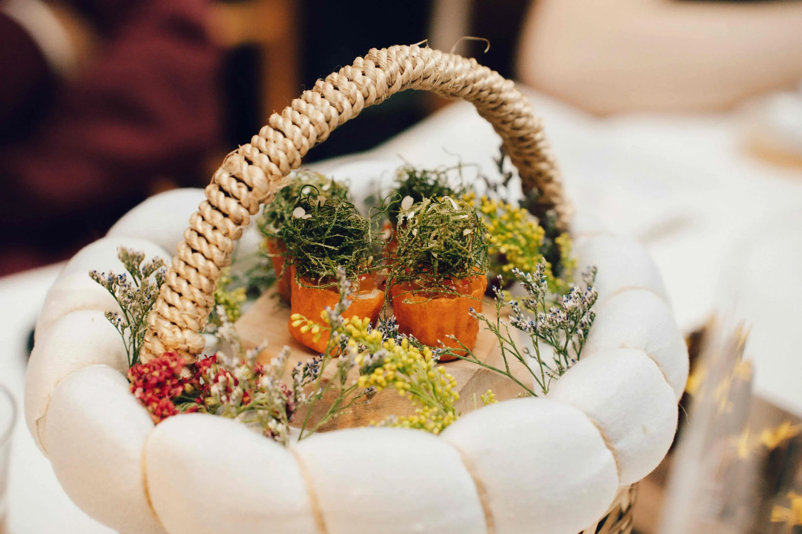 Close-up of a woven wicker basket handle cradling a bright floral arrangement with small yellow, purple, and green sprigs in white-gloved hands.