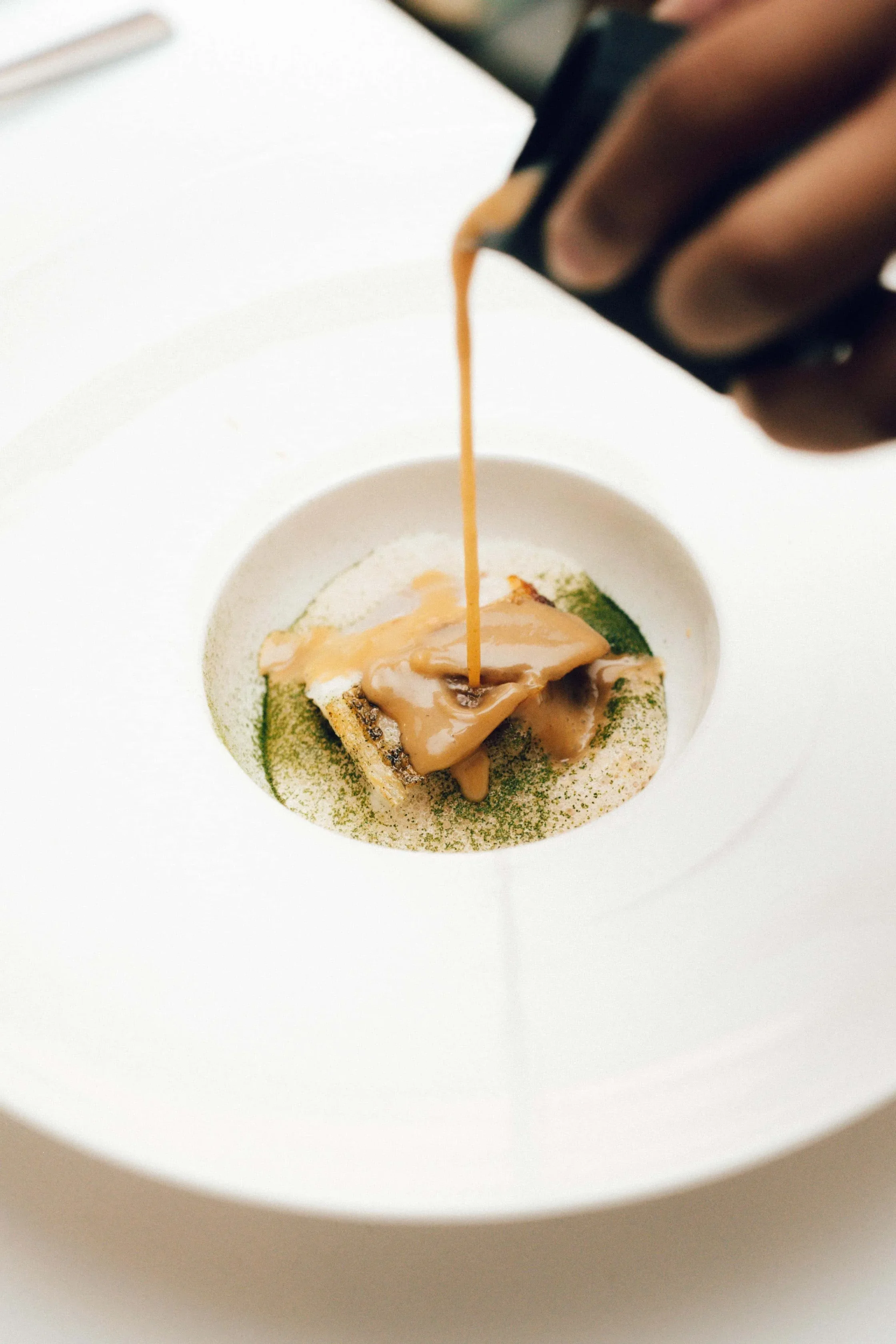 Person pouring orange sauce over a small round dessert with green powder on a white plate.