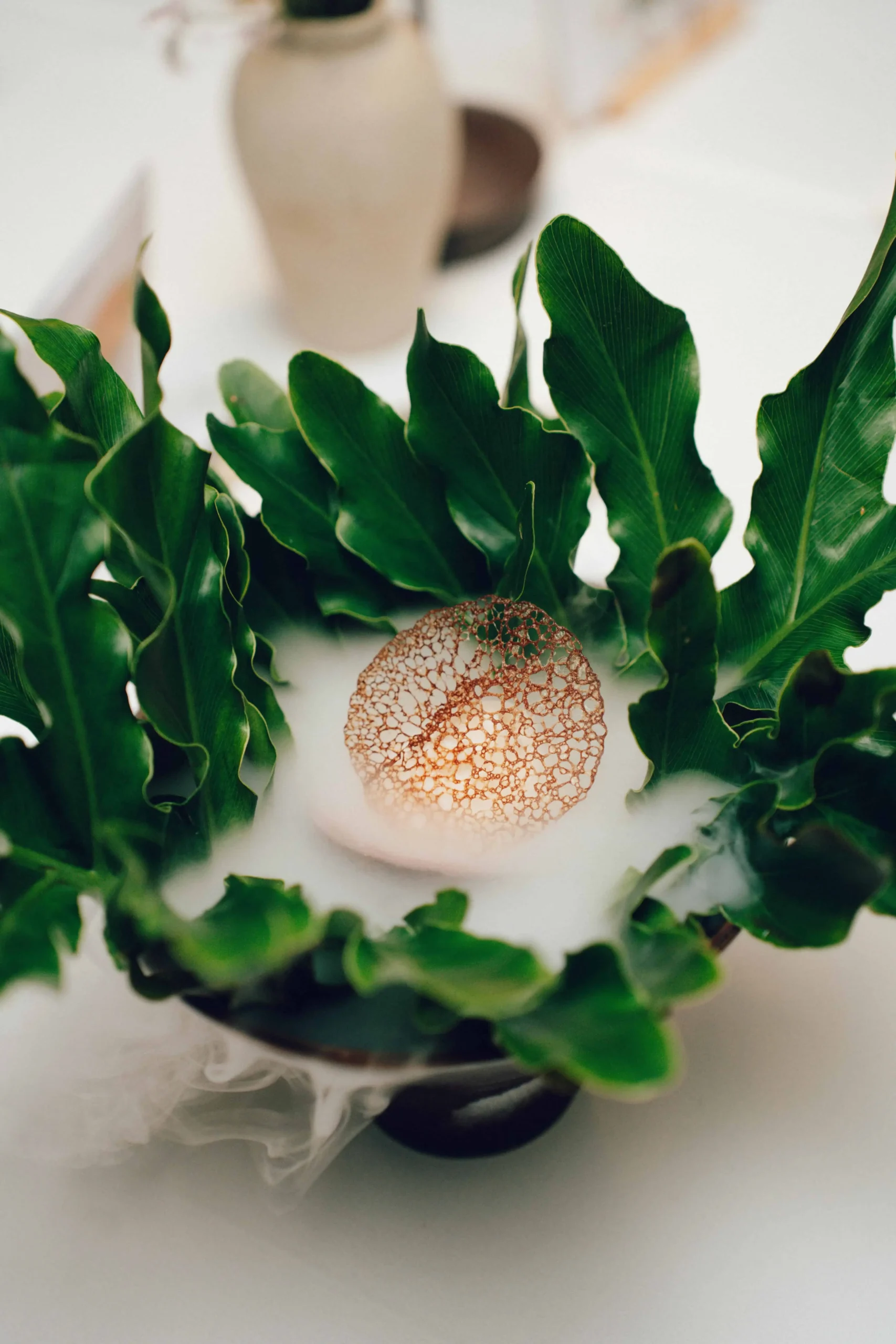 Green leafy plant arrangement in a white vase with a copper lattice ornament centerpiece.