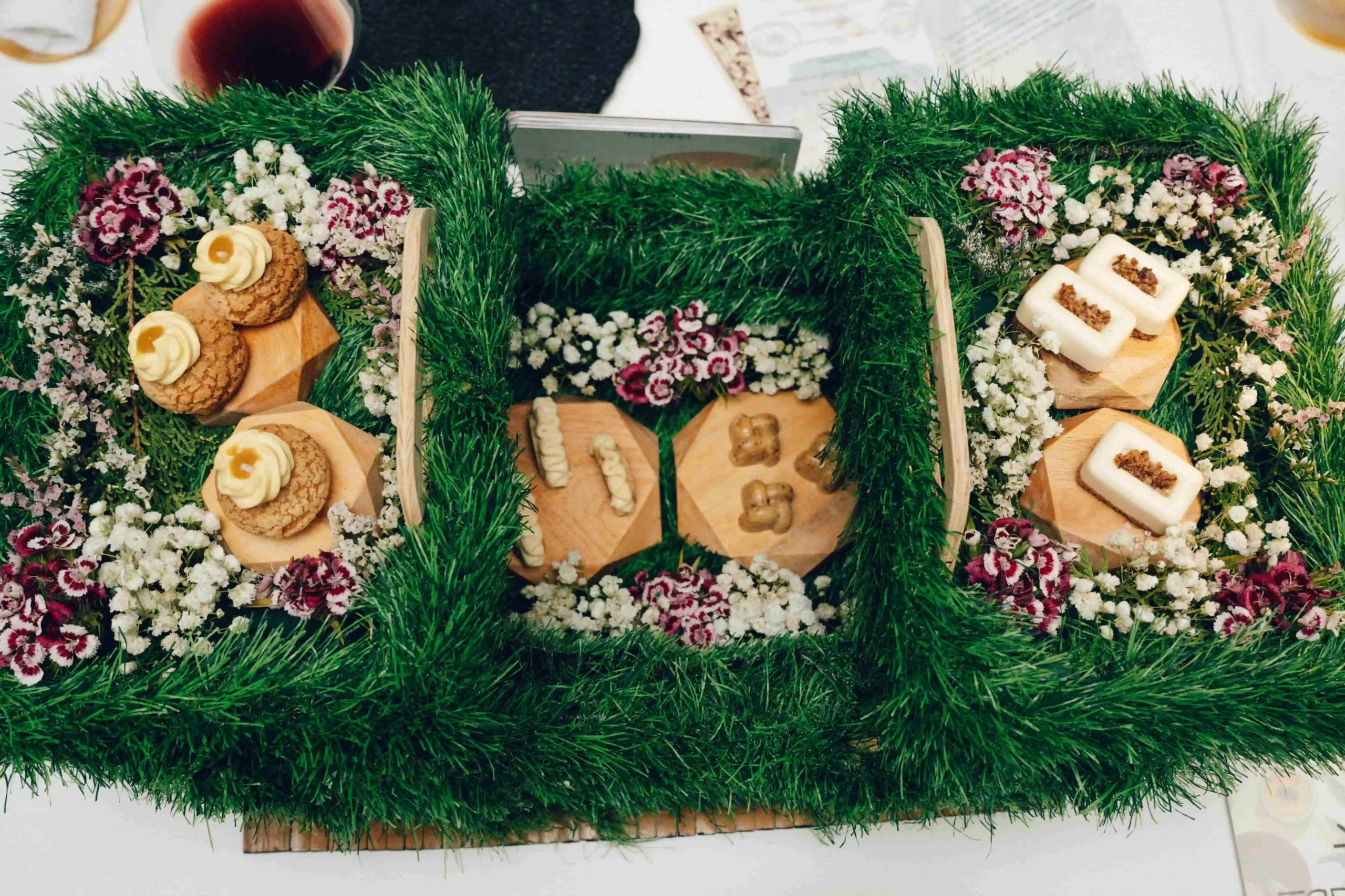 Top-down view of a festive display: green garland with small pastries and desserts arranged on wooden boards among white and red flowers.