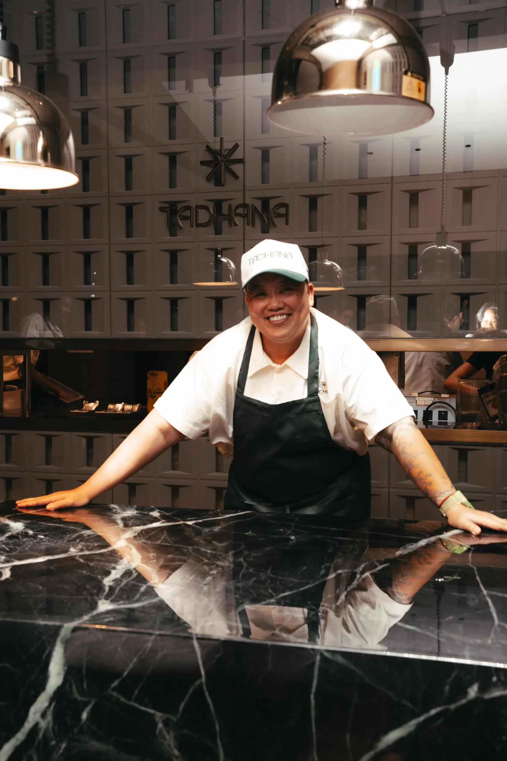 Smiling chef leaning on a dark marble counter in a white shirt and dark apron, with a Tadahana cap in a modern kitchen.