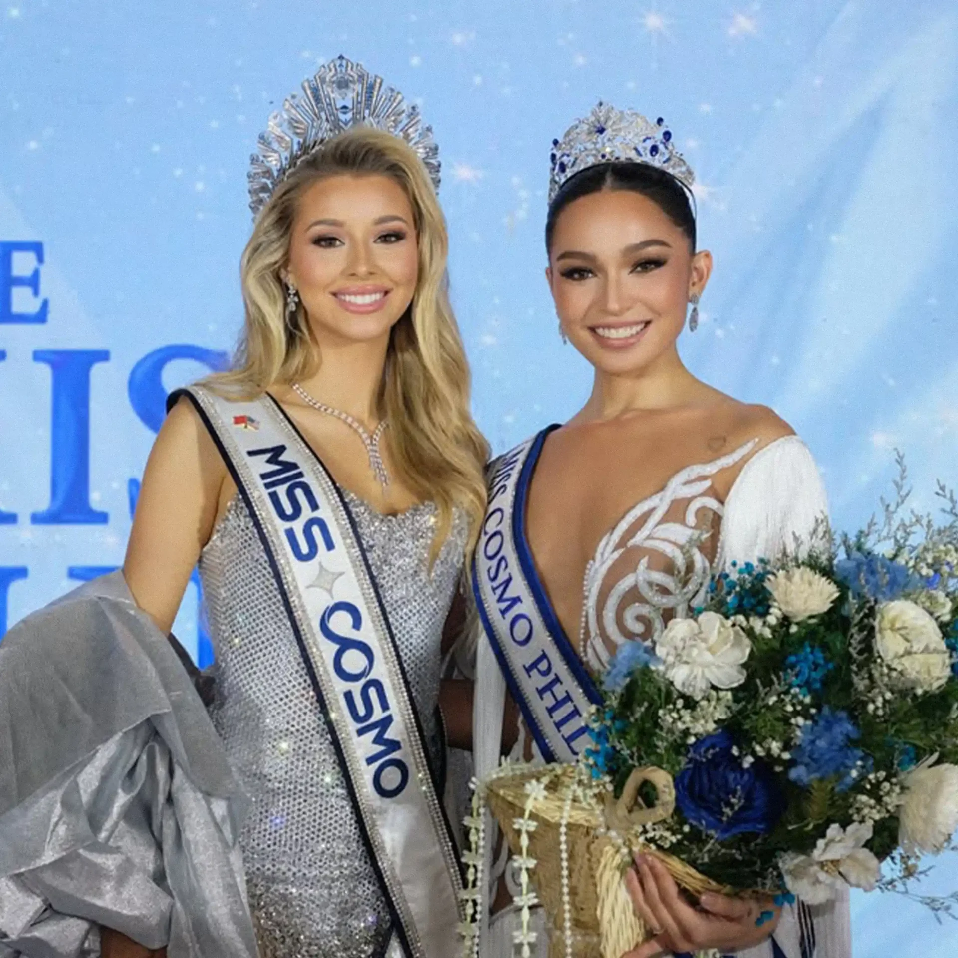 Two beauty queens in crowns and sashes posing together, holding a bouquet against a blue backdrop.