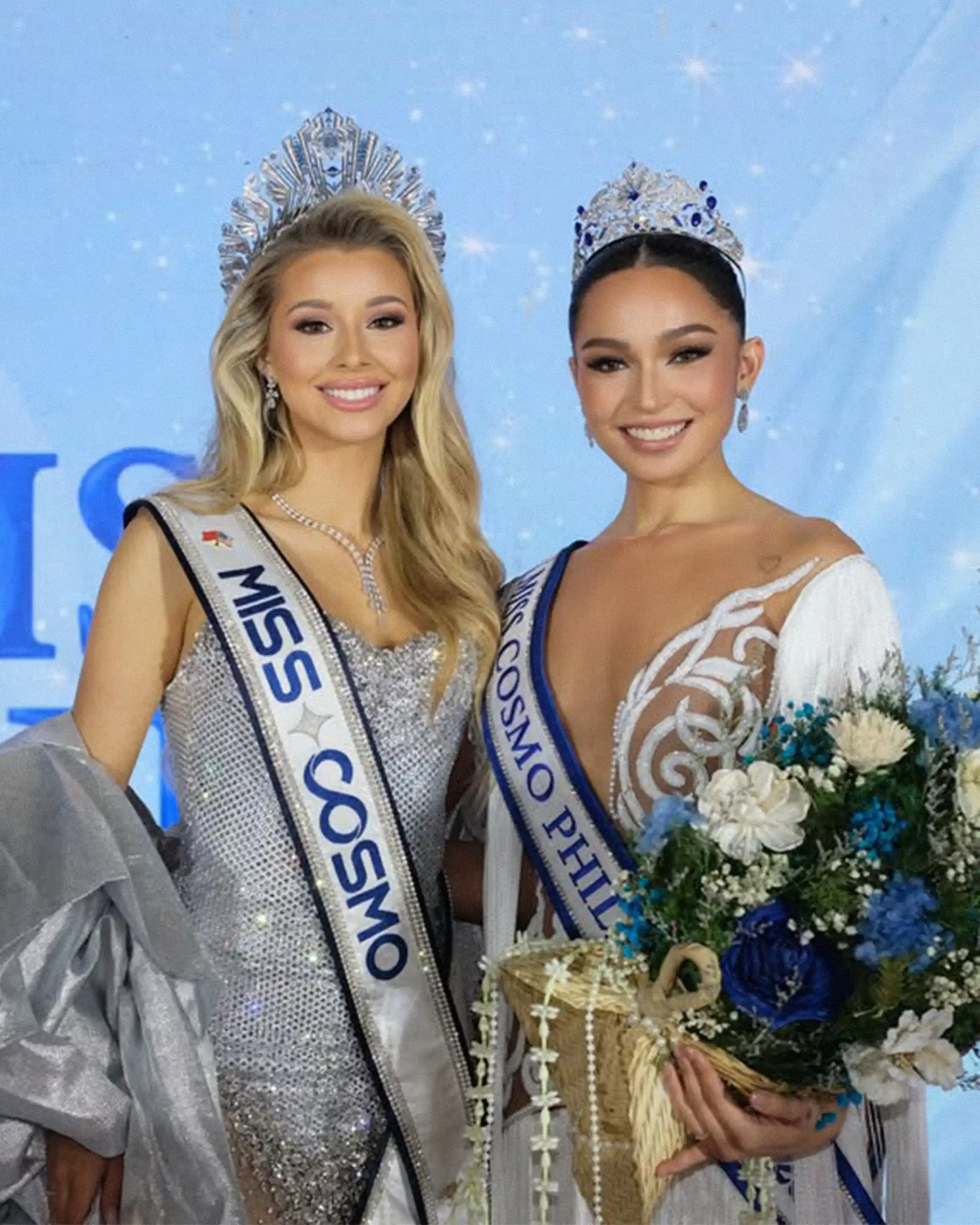 Two pageant contestants wearing crowns and sashes pose together, smiling; one holds a flower bouquet.