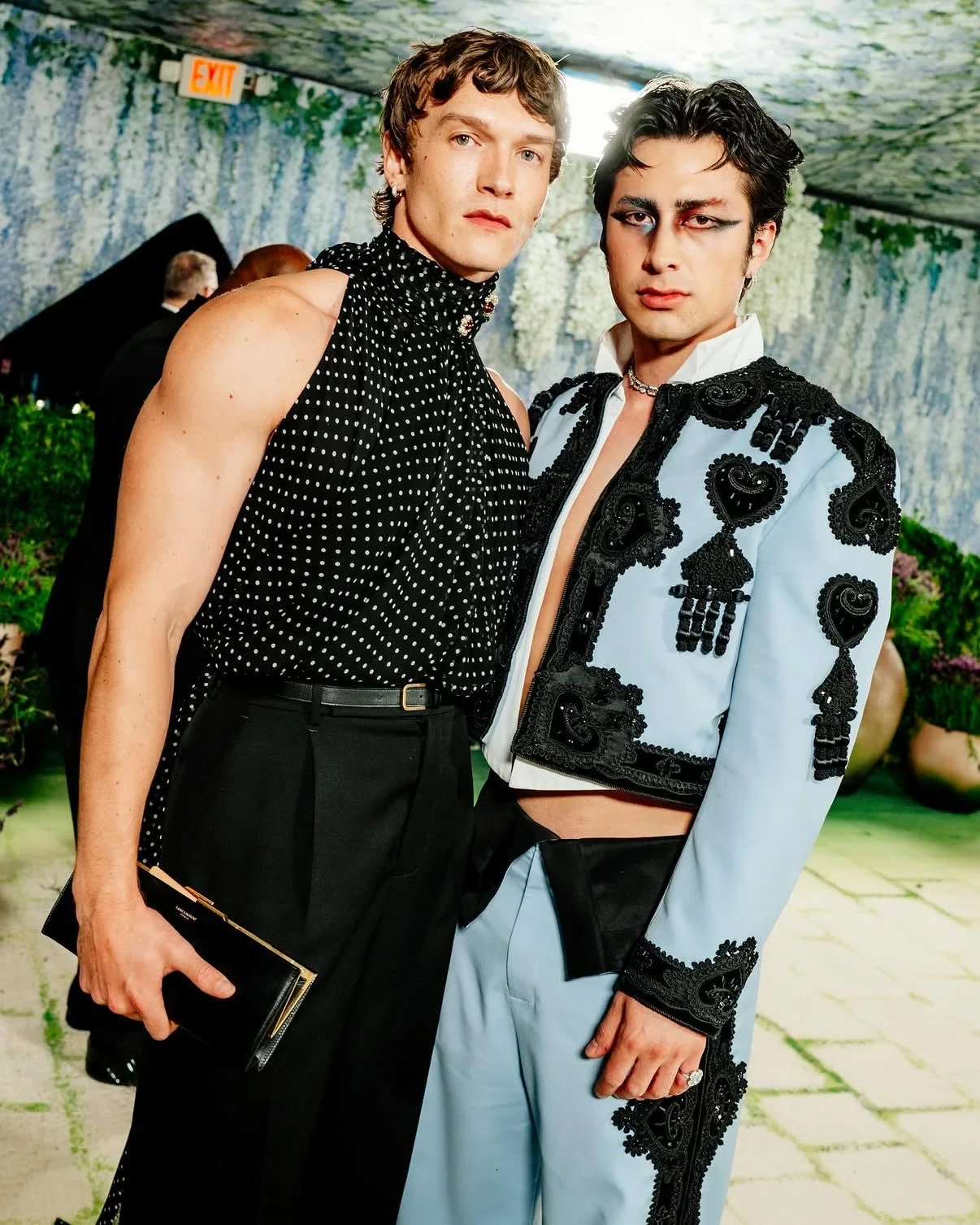 Two stylish men pose together at an event, one in a black polka-dot sleeveless top and black trousers, the other in a light-blue embellished suit with black lace details.