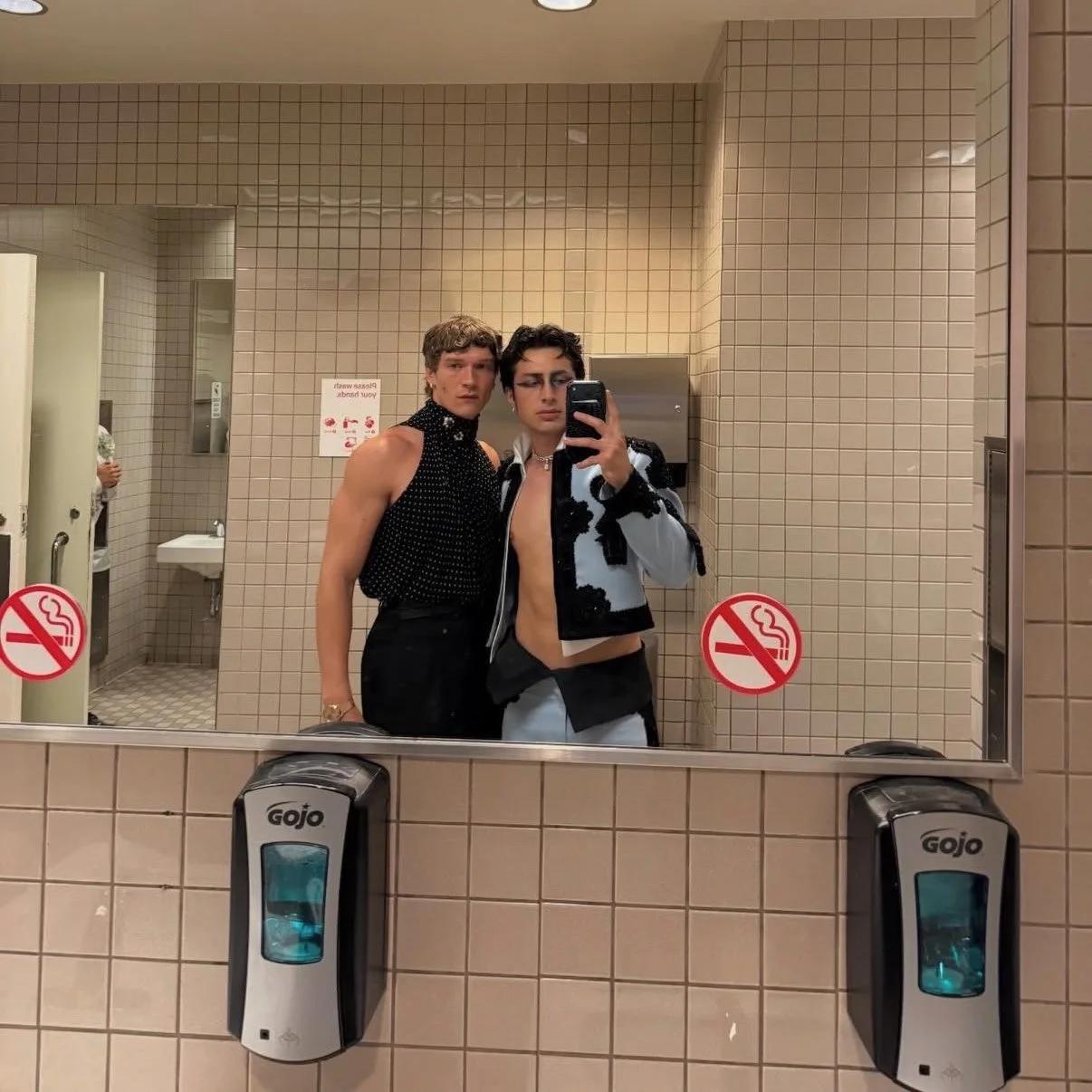 Two young men take a mirror selfie in a beige tiled bathroom; one shirtless with a black patterned vest, the other in a blue-and-black jacket.