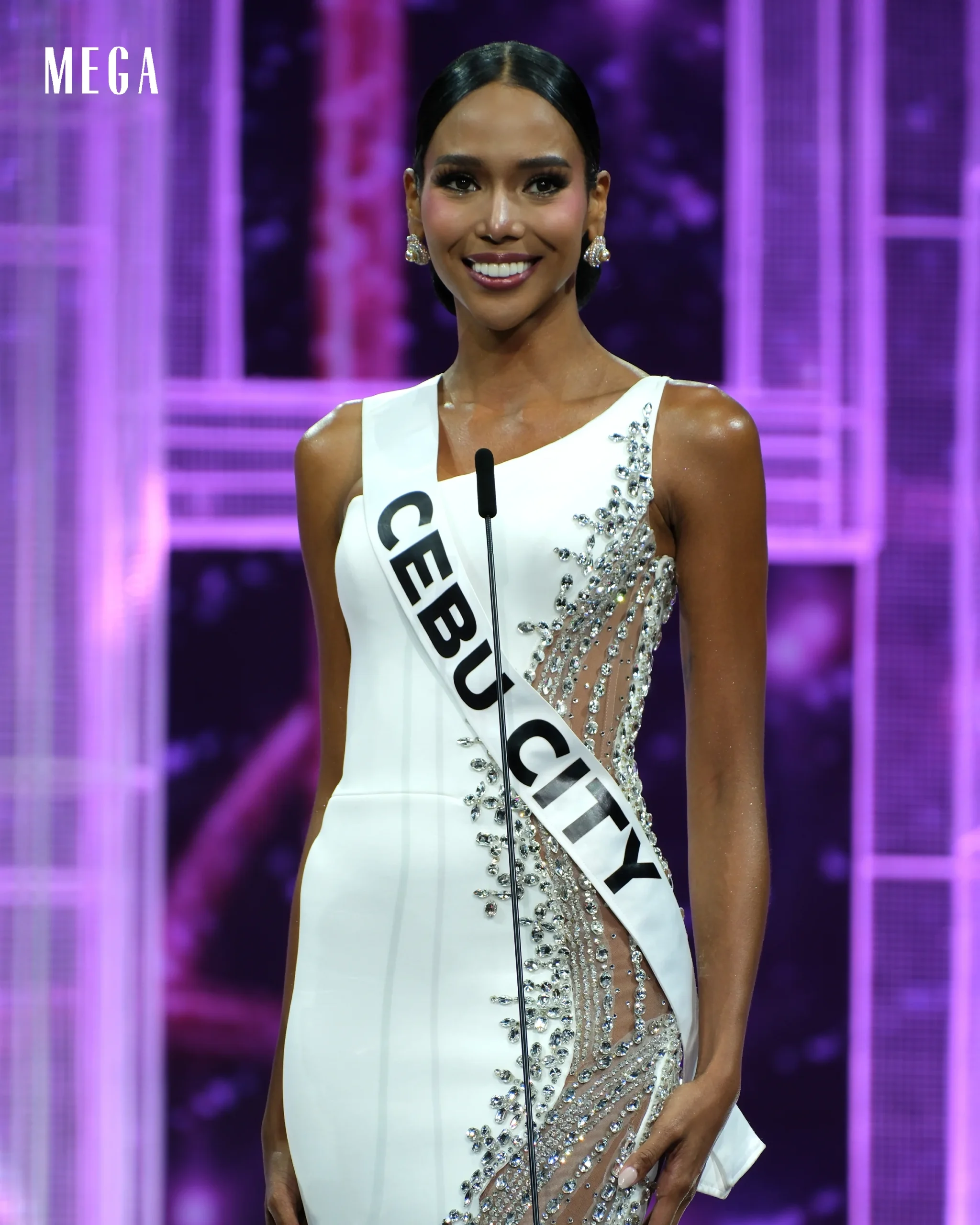 Miss Georgia contestant on stage in a white beaded gown with a sash reading 'GEORGIA', purple-lit background, smiling at the mic.