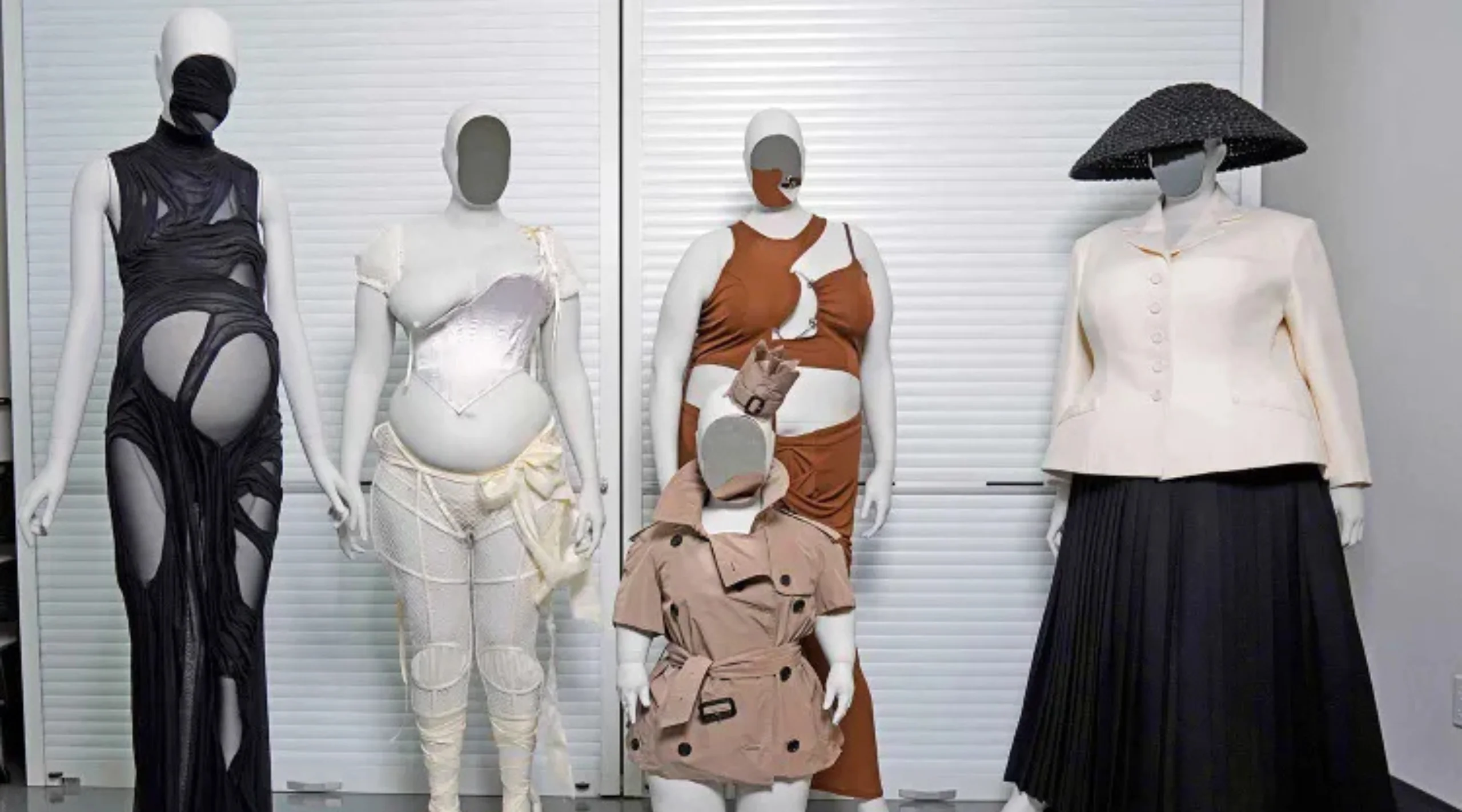 Four mannequins pose in a fashion showroom wearing avant-garde dresses: a black cut-out gown, a white corseted gown, a brown-and-beige asymmetric dress, and a cream blazer with a black pleated skirt with hat, against light panels.