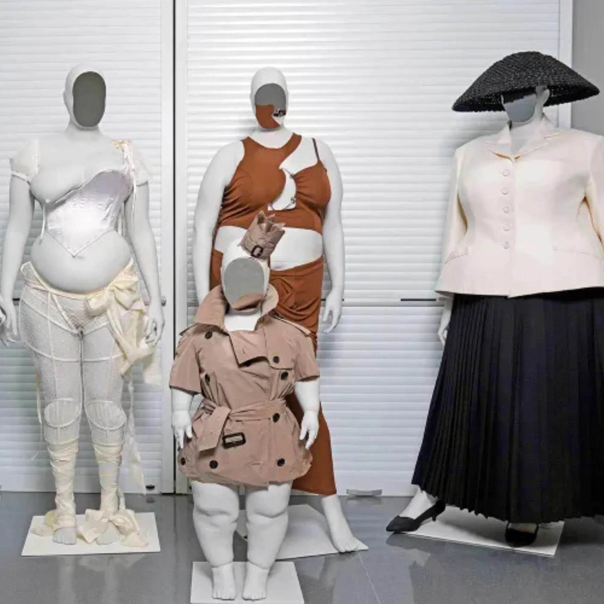 Three mannequins in a shop window wearing dresses: white gown (left), brown-and-white wrap dress (center), and a pale blouse with a large black hat (right).