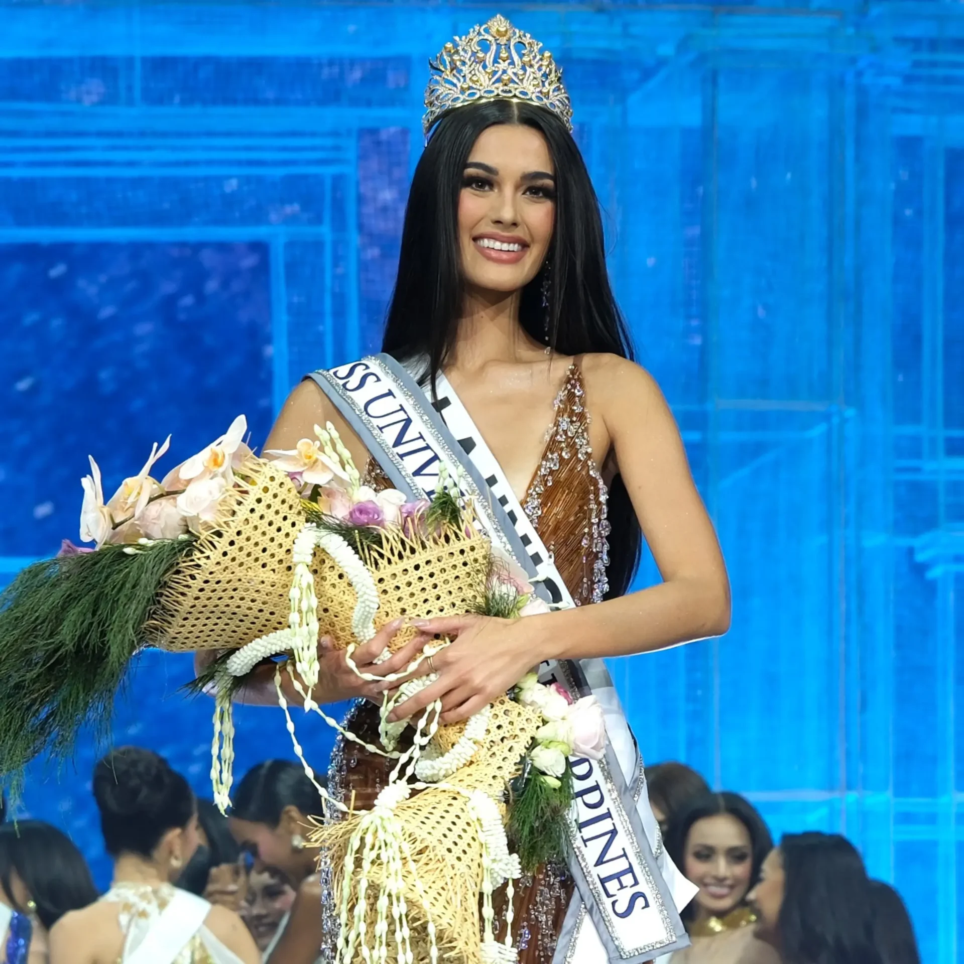 Beauty pageant contestant wearing a crown and sash, smiling while holding a large flower bouquet on a blue stage backdrop.