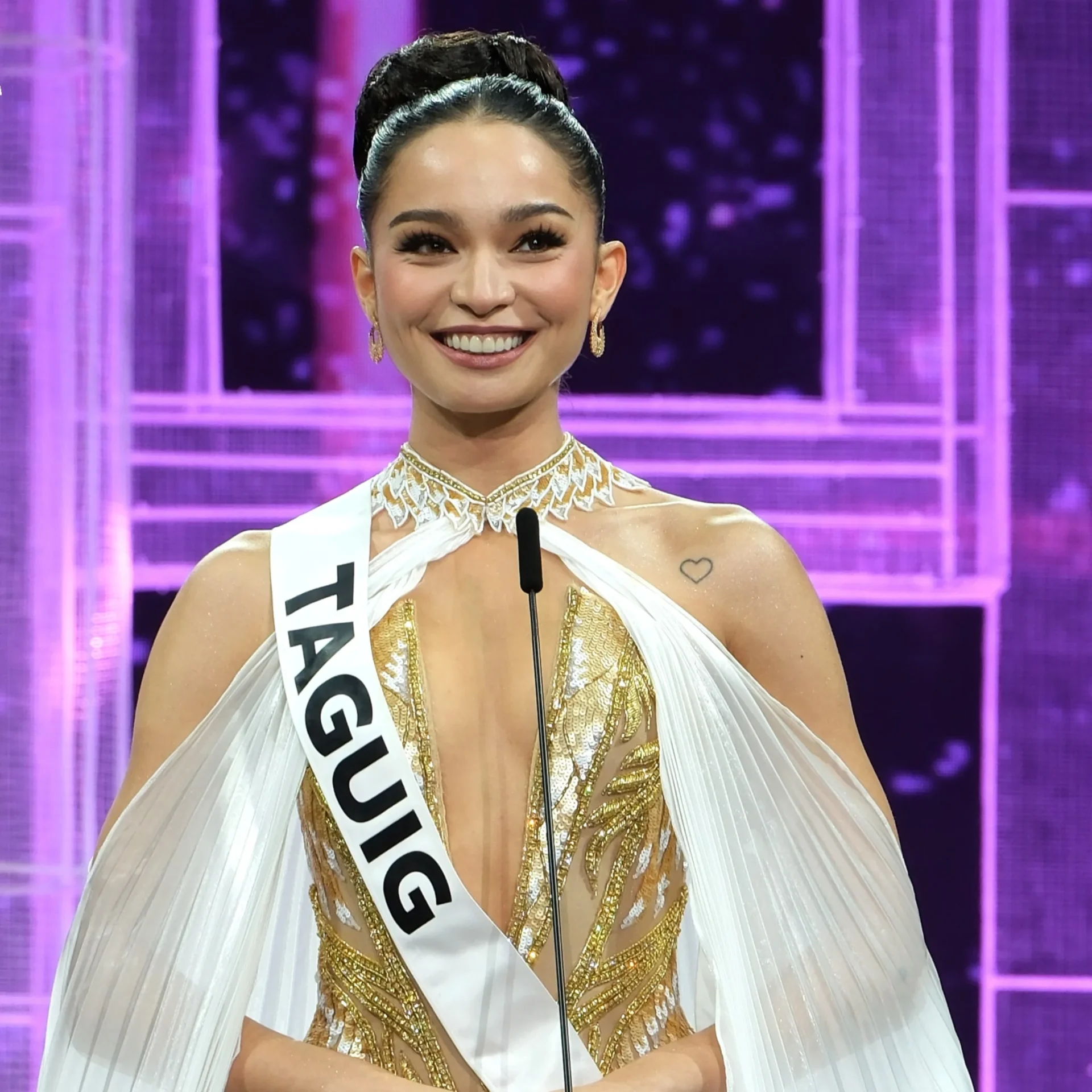 Smiling pageant contestant in a white-and-gold gown with a sash, standing at a microphone on a purple-lit stage.