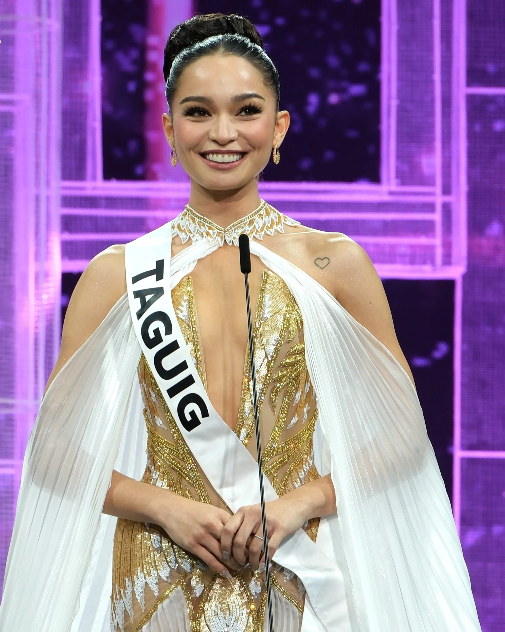 Smiling pageant contestant in a gold sequined gown with a white cape, standing at a microphone on a purple-lit stage.