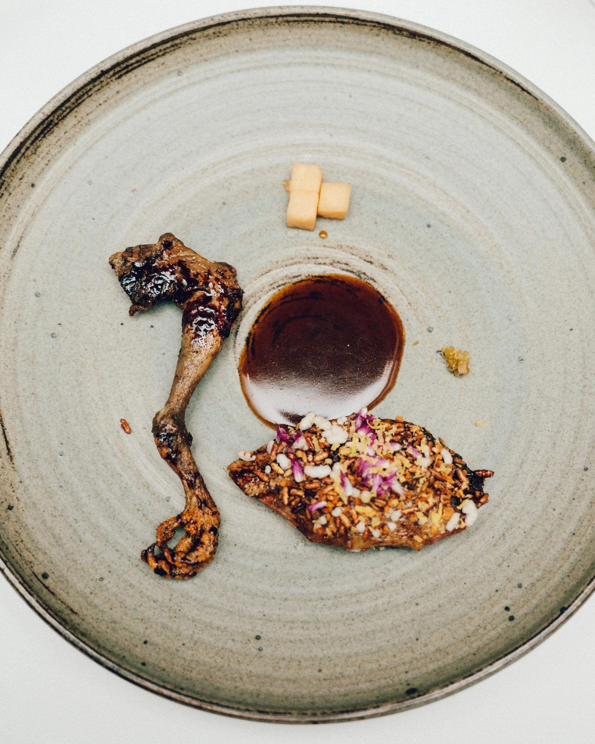 Ceramic plate holding a fried chicken leg, a small pool of brown sauce, a crumb-coated meat piece topped with nuts, and three small cheese cubes nearby.