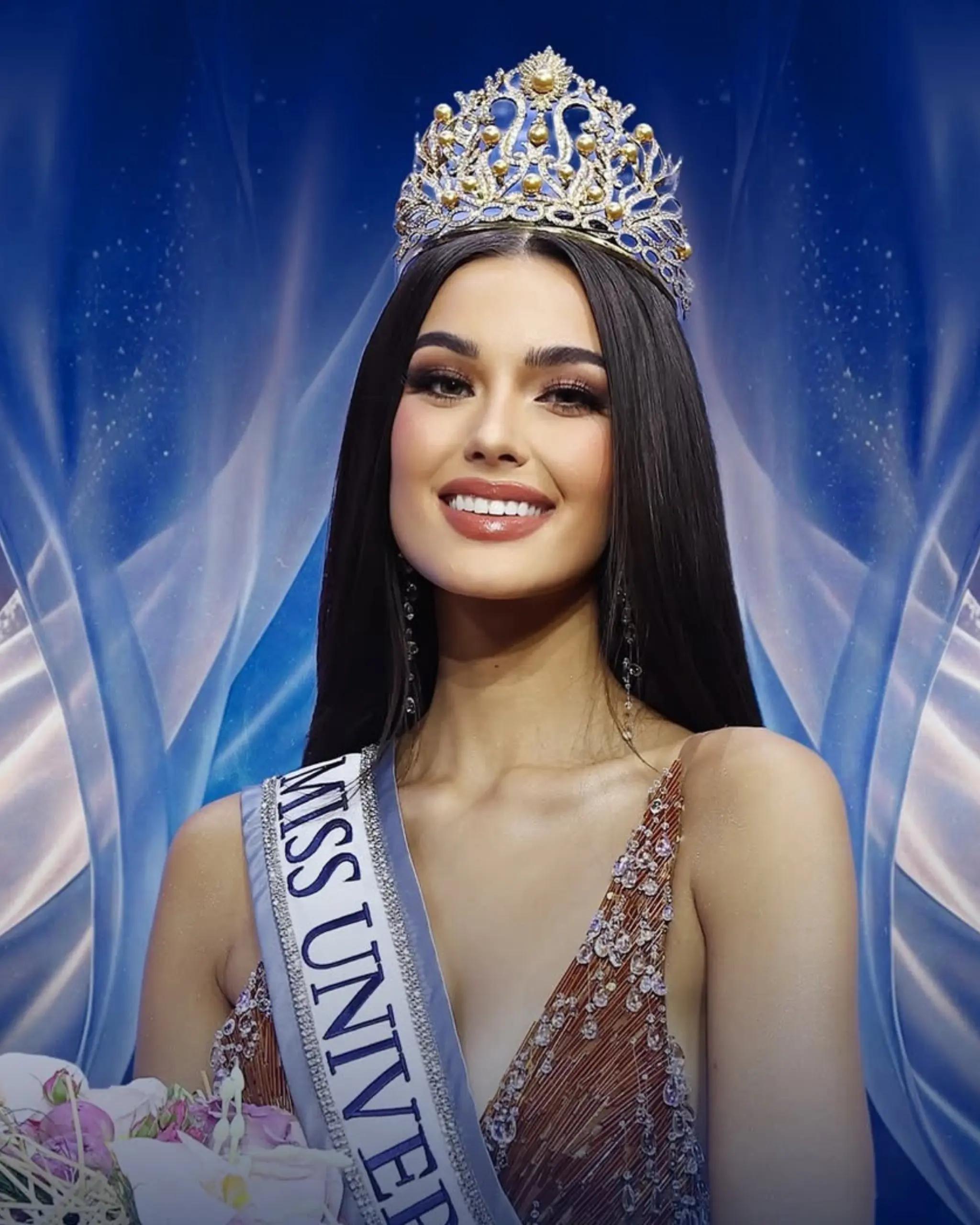 Beauty queen wearing a jeweled crown and sparkling gown, smiling on a blue stage backdrop with a bouquet nearby.