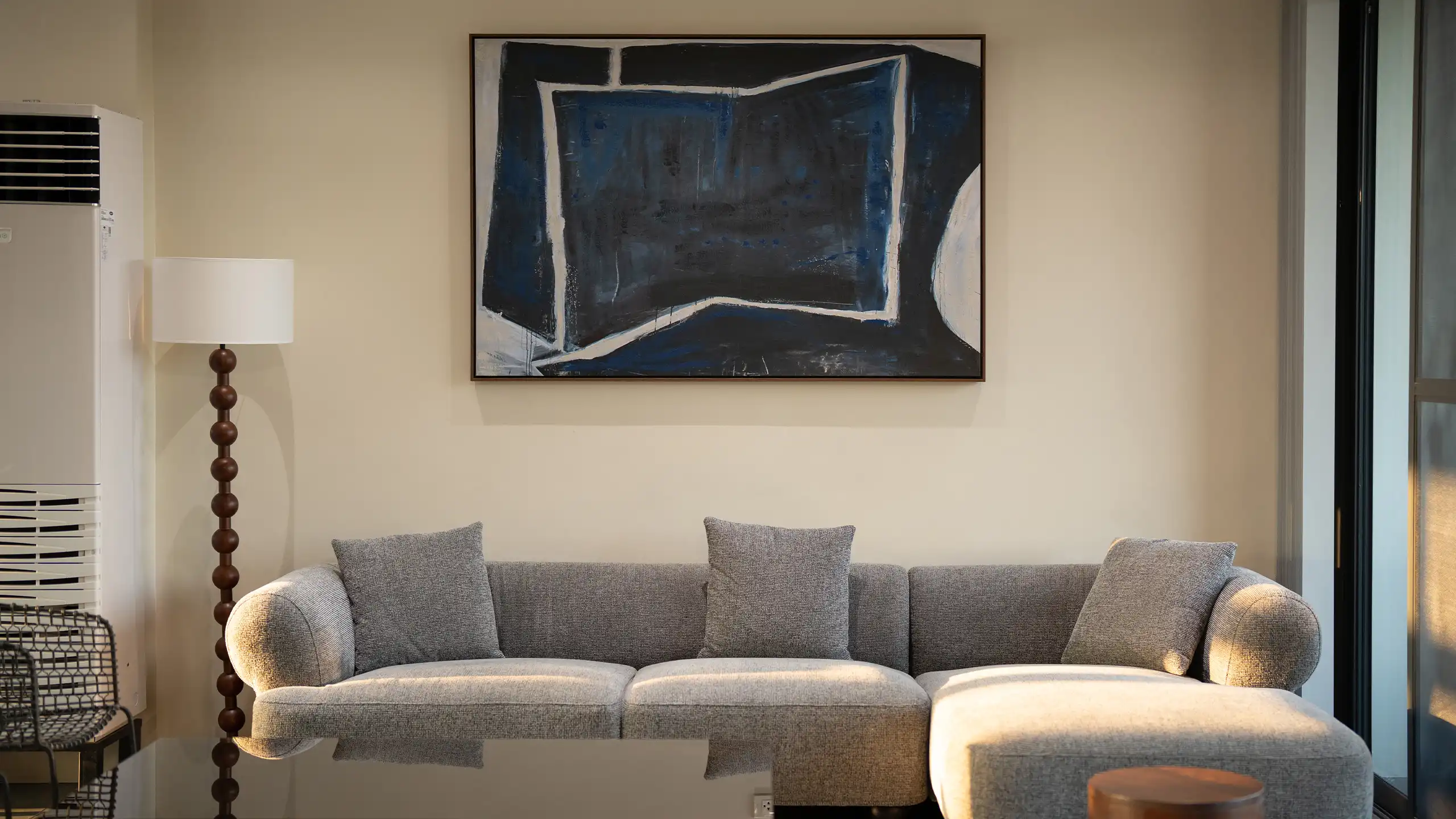 Living room scene with a grey sectional sofa, abstract blue-black painting, and a tall wooden floor lamp beside an air conditioner unit on the left.