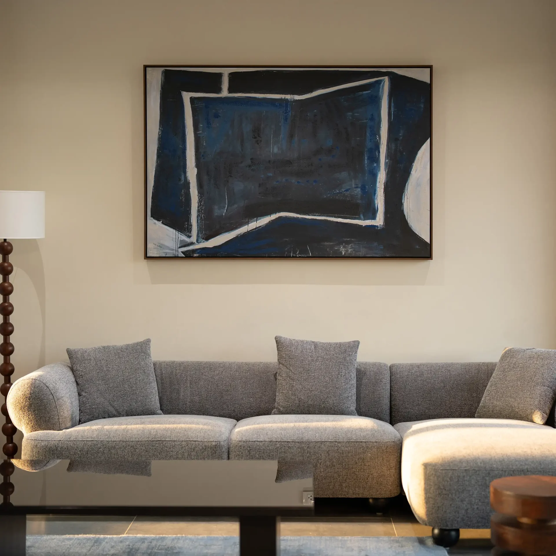 Abstract blue-gray geometric painting hung on a beige wall above a plush gray sofa in a living room.