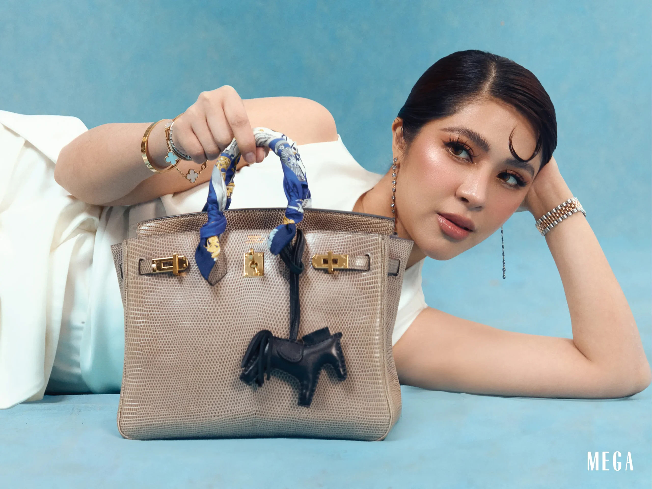 A fashion shoot close-up of a woman posing with a textured beige handbag, scarf tied to the handle, against a blue background.