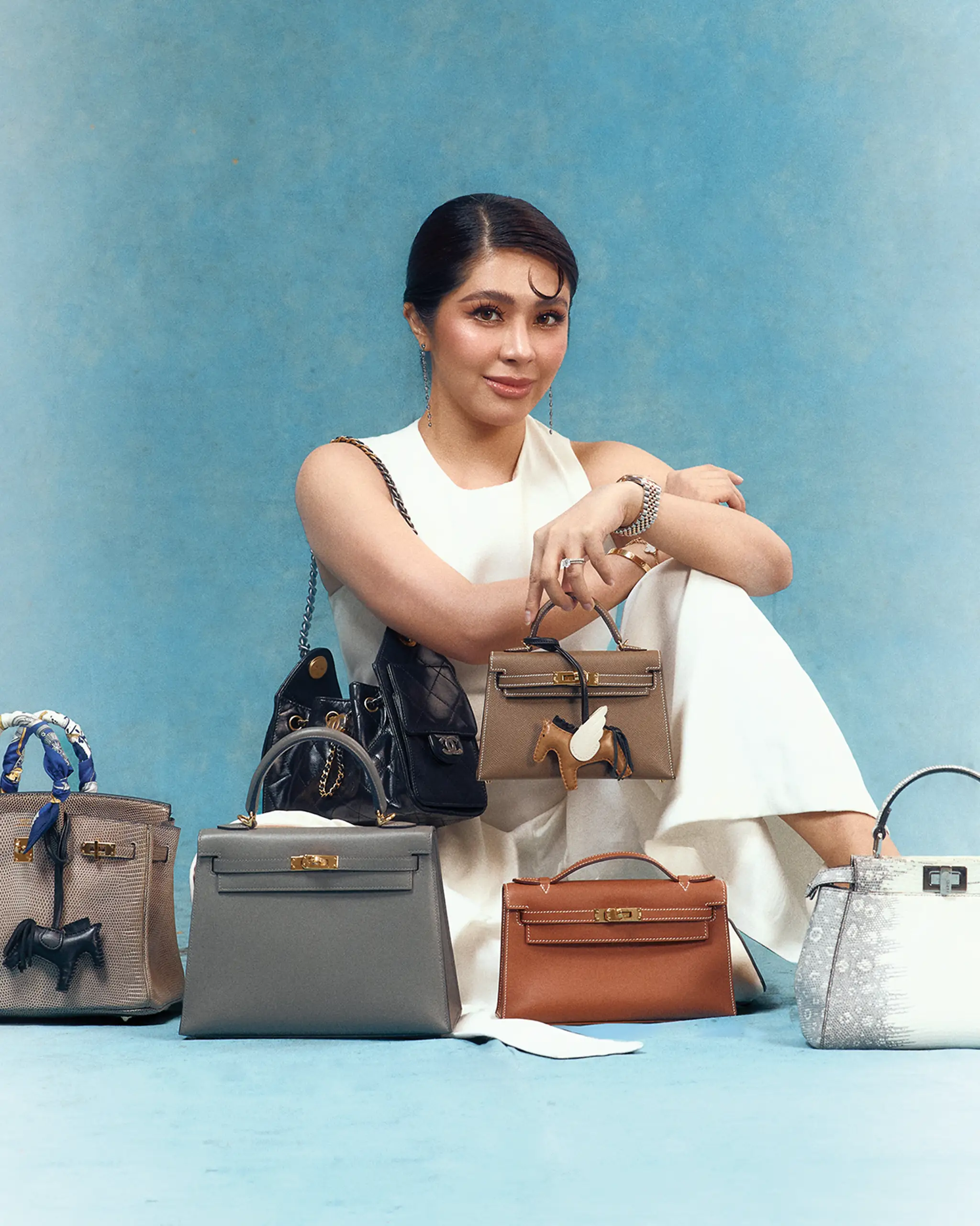 Fashionable woman in white, posing with a variety of handbags arranged around her against a blue backdrop.
