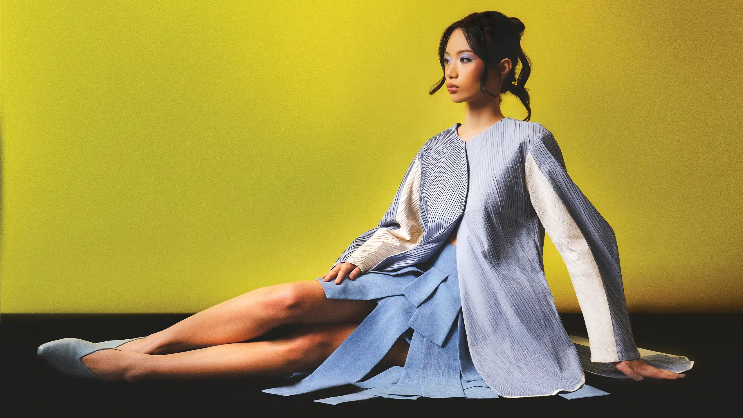 Model seated on the floor against a bright yellow backdrop, wearing a light blue dress with a blue-gray striped cape sculpting the silhouette.