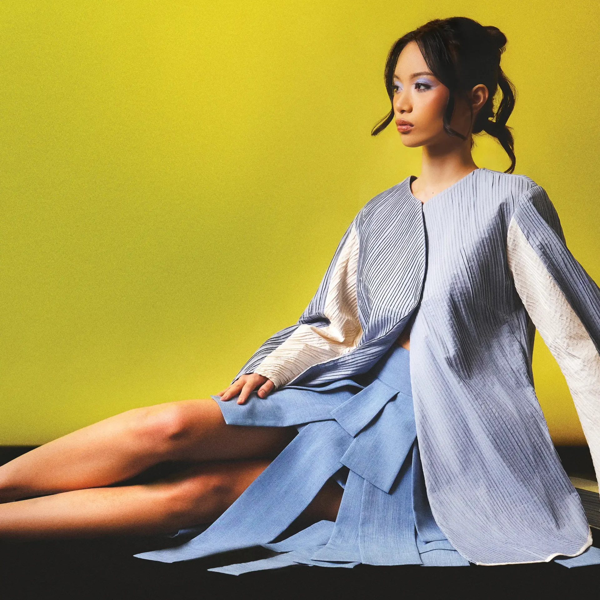 Woman seated in a flowing blue-striped kimono-like robe, legs extended forward, against a bright yellow background.