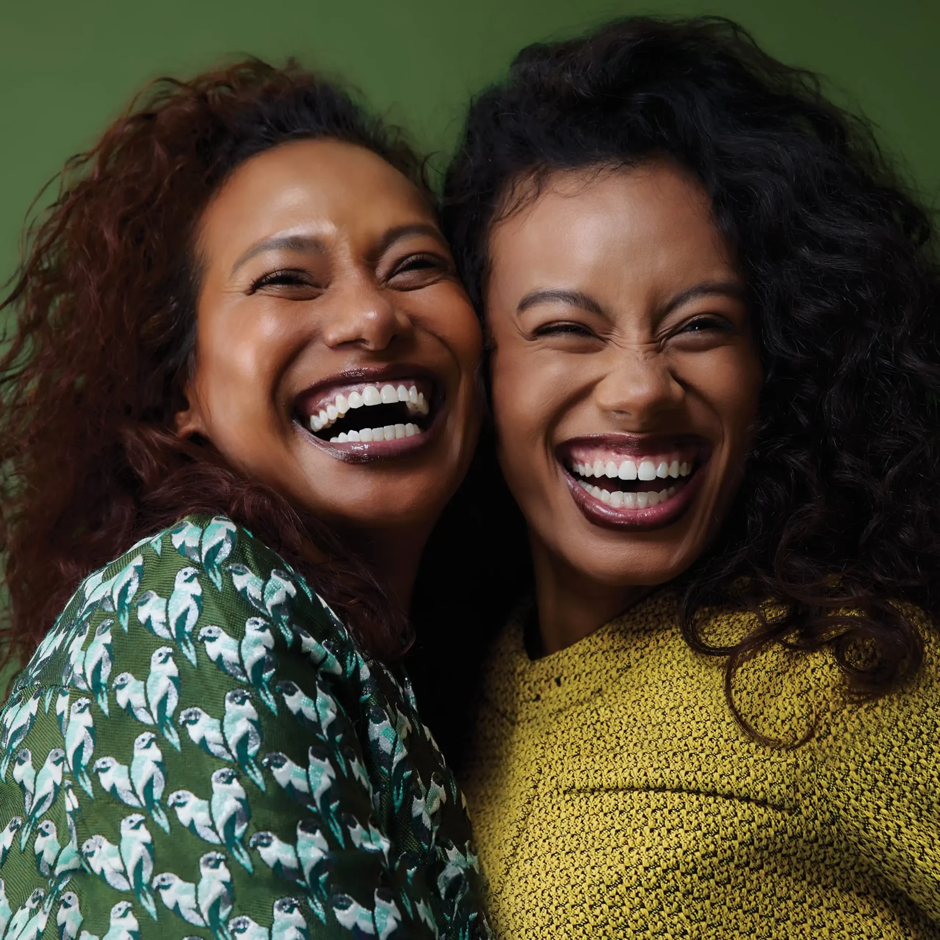 Two women smiling broadly, close together, against a green background.