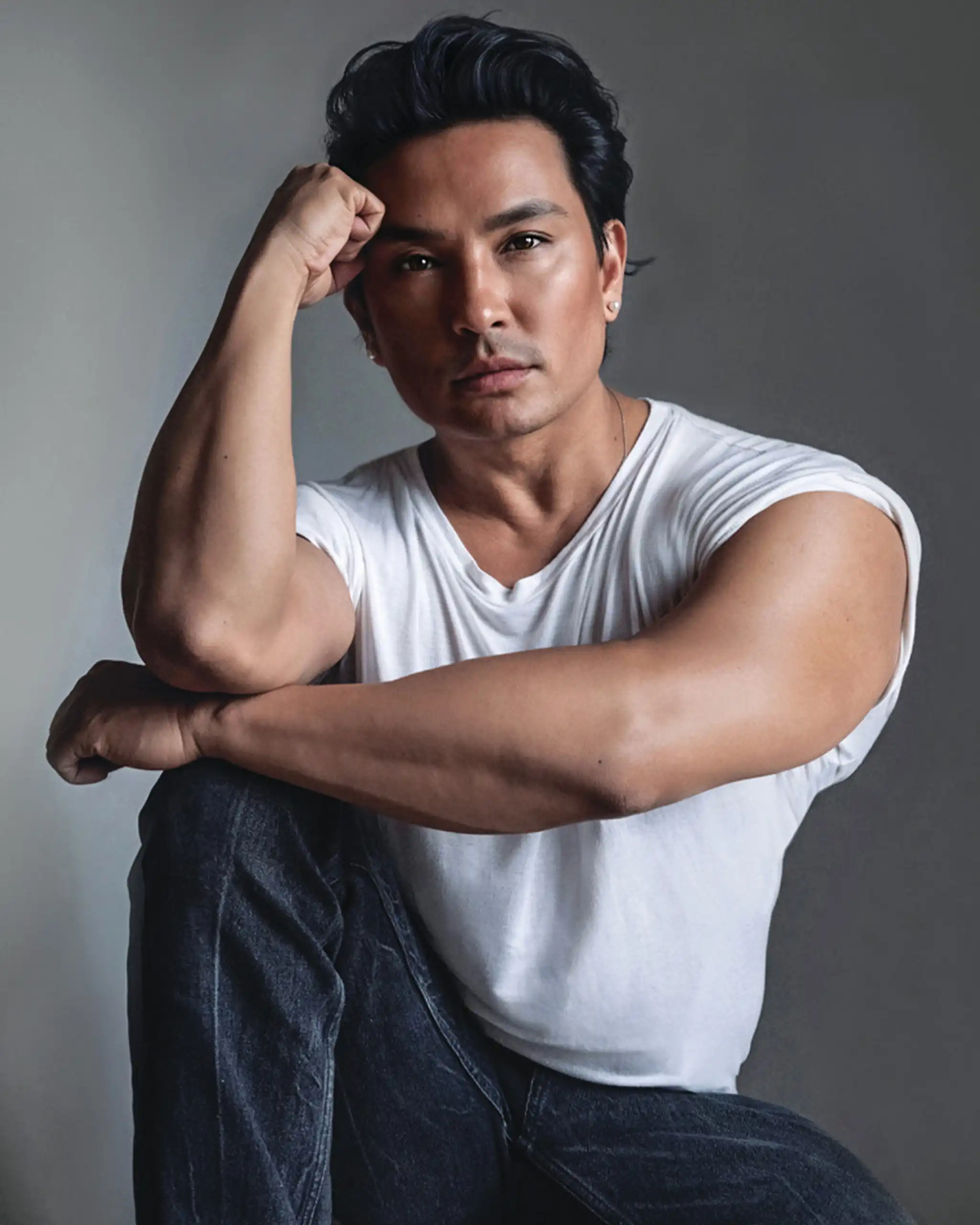 Man posing thoughtfully with his elbow on a knee, hand supporting his head, wearing a white t-shirt and jeans in a neutral studio backdrop.