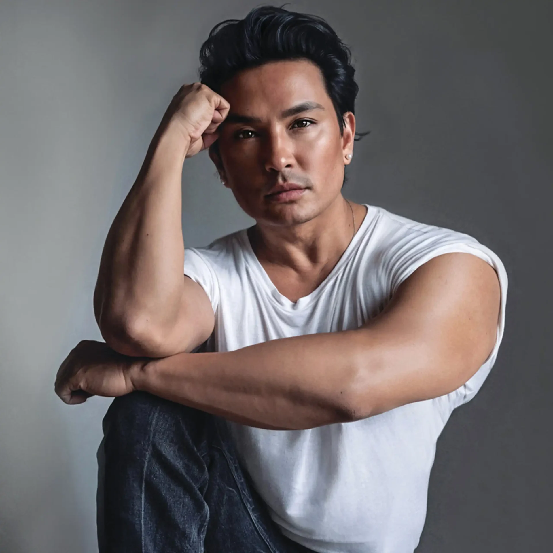 Man with dark hair wearing a white shirt, resting his head on his hand in a thoughtful pose against a gray backdrop.