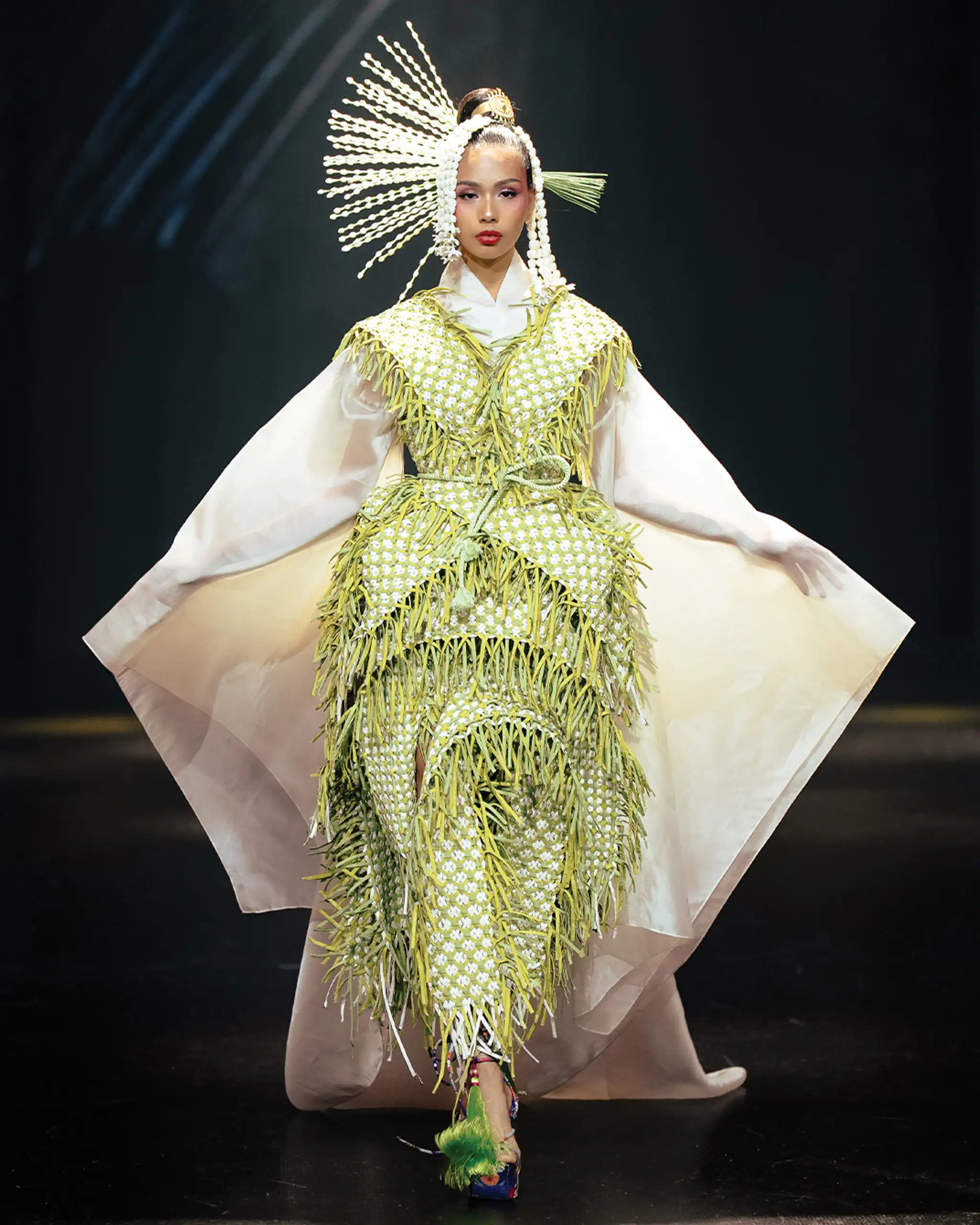 Model on a runway wearing a green fringed gown with a dramatic white headdress and long white sleeves.
