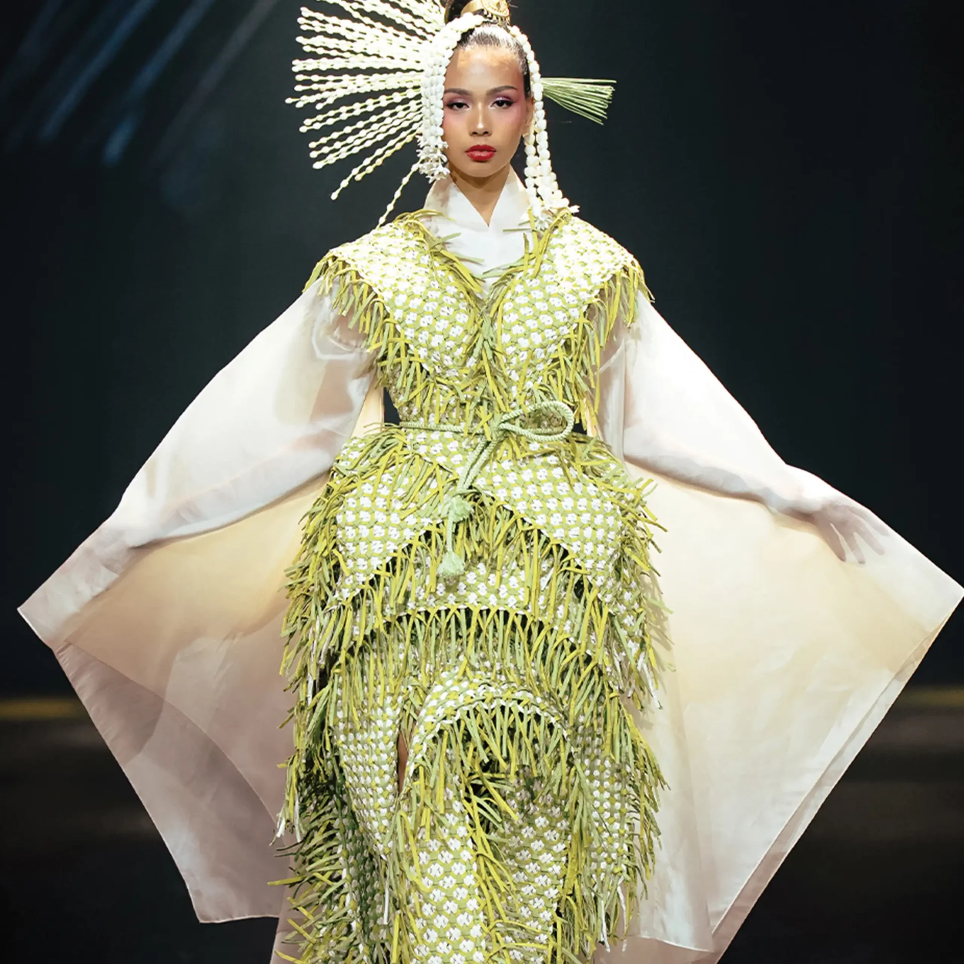 Model on a runway wearing a pale dress with a yellow floral fringe cloak and a large white shell headpiece against a dark background.