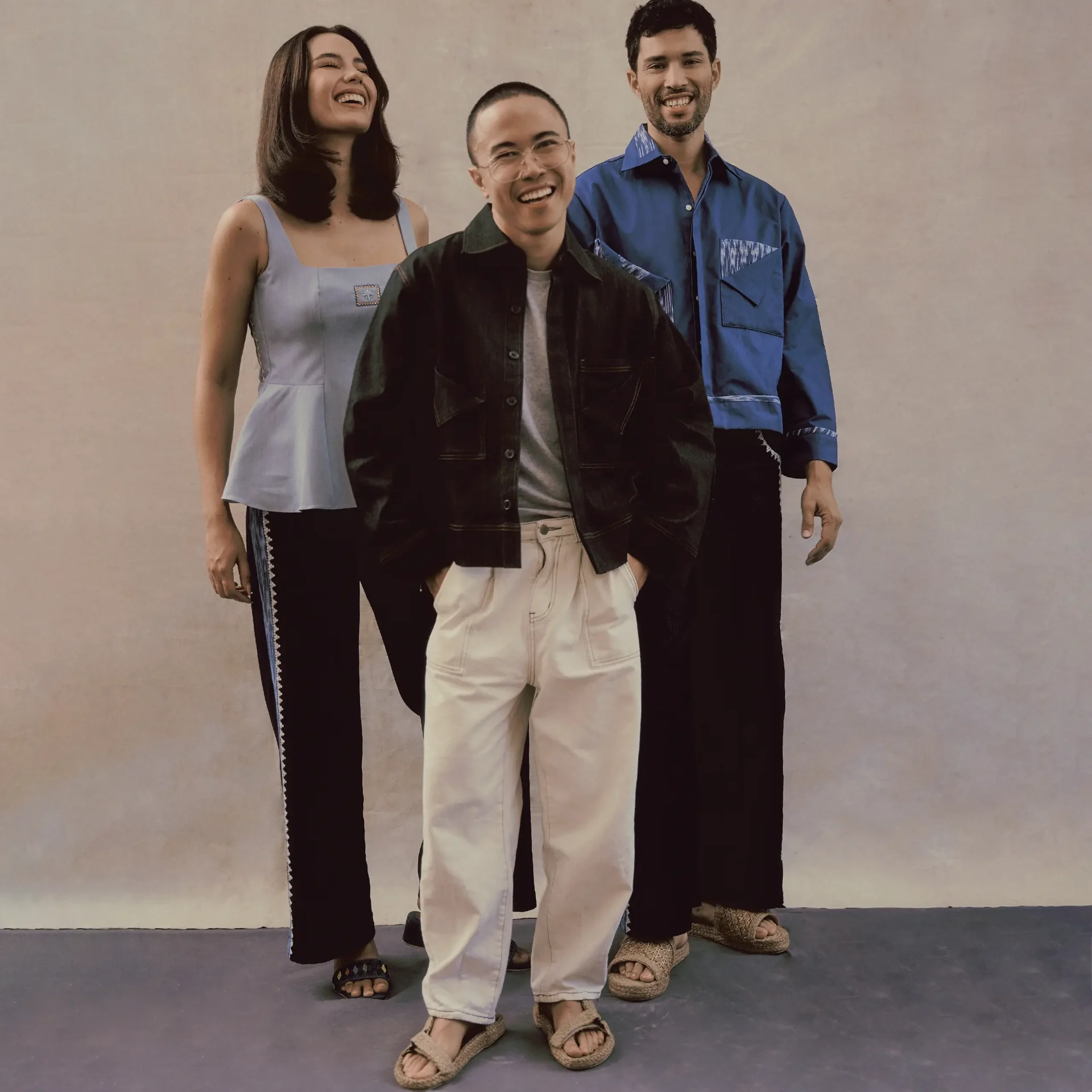 Three friends standing close together and smiling, wearing casual outfits and sandals in front of a neutral backdrop.