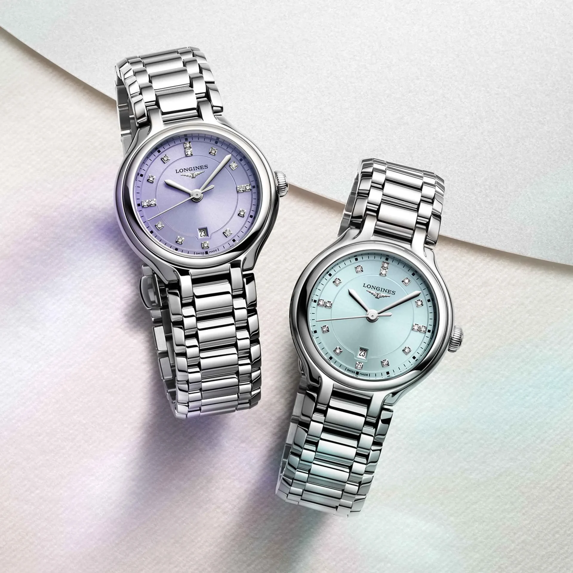 Close-up of three silver wristwatches with pastel lavender and light blue dials on a soft fabric background.