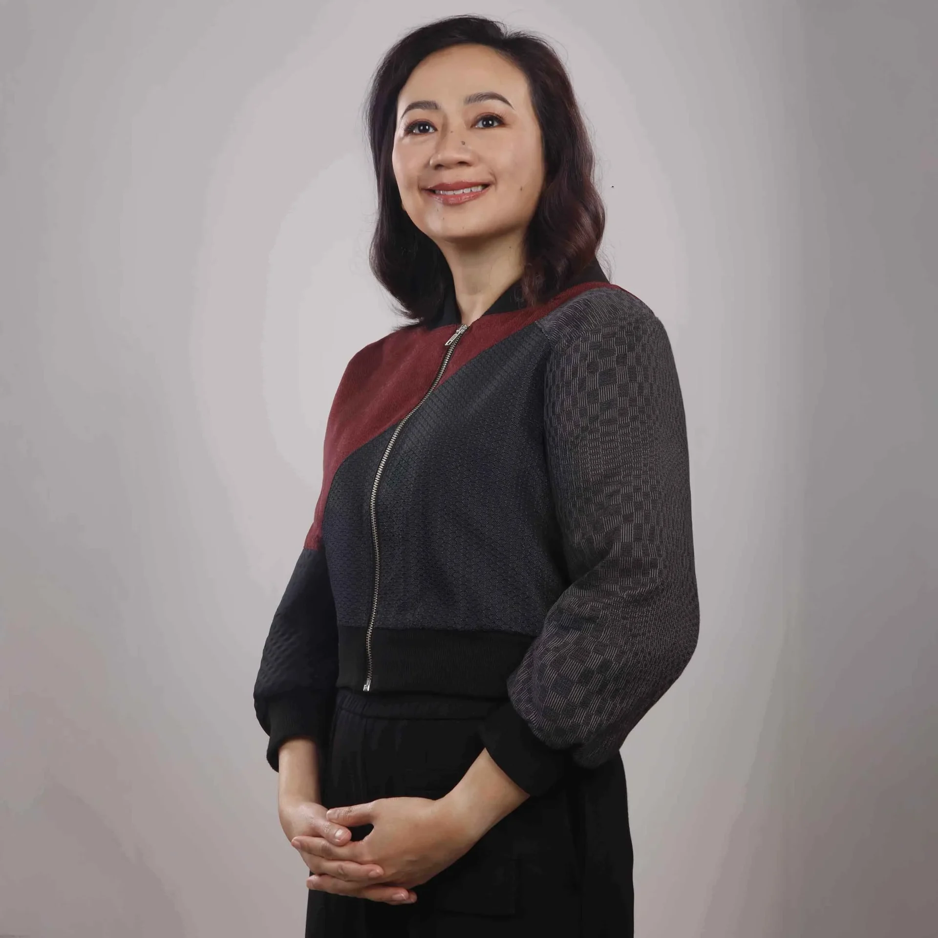 Woman standing with arms crossed, wearing a dark color‑block cardigan against a gray studio backdrop.